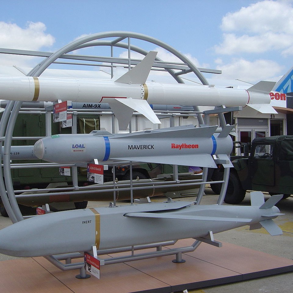 L'image montre une exposition de missiles sur un socle. On peut voir plusieurs types de missiles, dont un missile de croisière, un missile air-air et un missile guidé. Les missiles sont présentés sur un support circulaire, avec des informations en dessous de chacun d'eux. En arrière-plan, on aperçoit des véhicules militaires et un ciel partiellement nuageux. C'est une scène typique d'une foire ou d'une exposition de défense militaire.