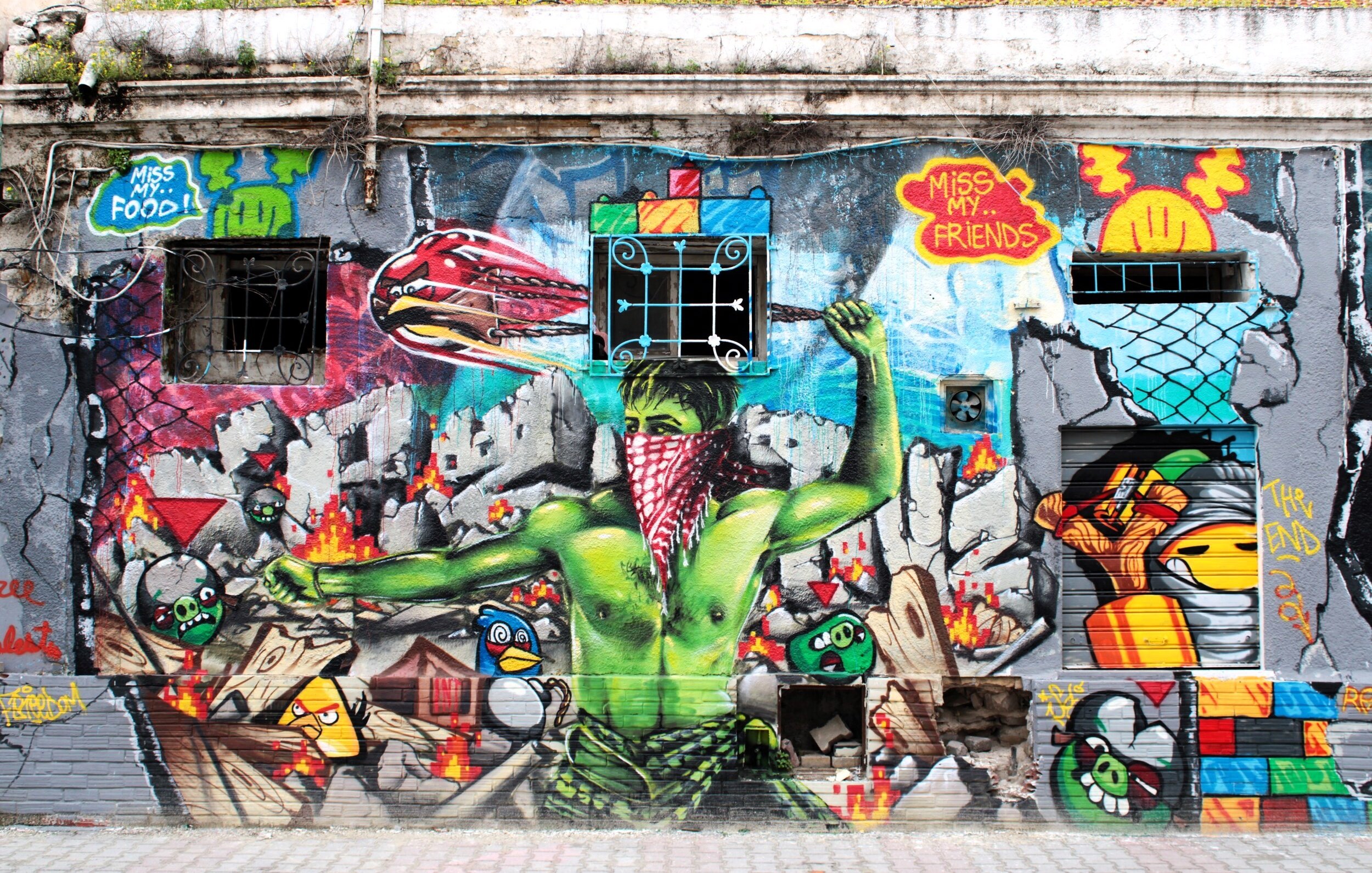 L'immagine mostra un murales colorato su un muro esterno. Al centro, c'è un personaggio verde con un fazzoletto rosso, che sembra esprimere un forte senso di protesta o di resistenza. Sullo sfondo, si possono notare elementi di graffiti e illustrazioni vivaci, tra cui simboli e oggetti in movimento, che creano un'atmosfera dinamica e urbana. Il murales combina diverse tecniche artistiche, rendendolo molto interessante visivamente.