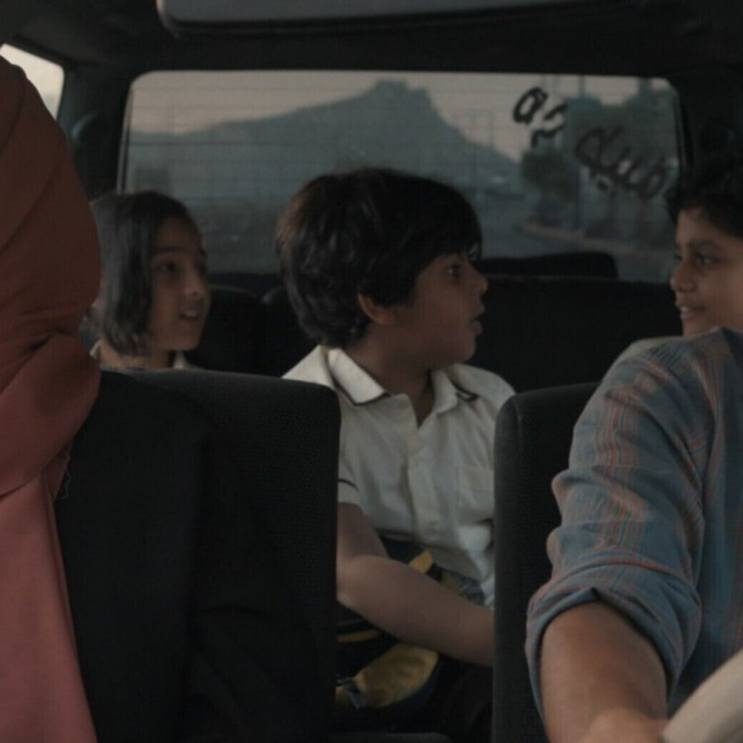 L'image montre un groupe de personnes dans une voiture. À l'avant, un homme au visage sérieux conduit, tandis qu'une femme portant un hijab est assise à côté de lui, semblant pensive. À l'arrière, deux enfants et un adolescent se parlent, créant une atmosphère de dynamisme et d'interaction. La lumière semble douce, suggérant une scène de fin de journée. L'ambiance générale évoque une sorte de voyage ou de déplacement, avec un mélange d'émotions au sein de la voiture.