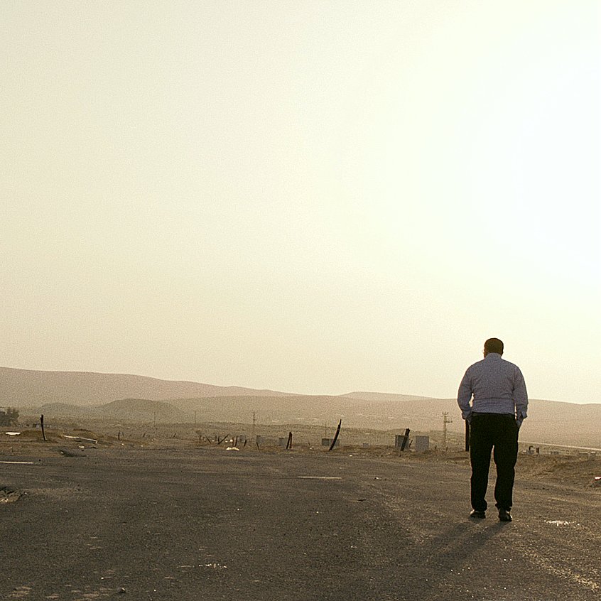 L'image montre un homme marchant seul sur une route déserte. On peut voir en arrière-plan des bâtiments modernes qui semblent inachevés ou abandonnés, avec un paysage désertique s'étendant à l'horizon. La lumière du soleil semble douce, suggérant une ambiance calme et un peu mélancolique. Le ciel est clair, et l'ensemble de la scène évoque une sensation de solitude et d'isolement.