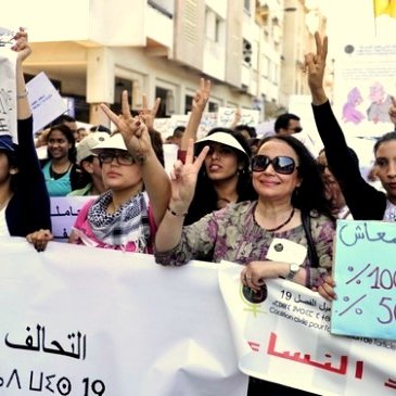 L'image montre un groupe de femmes participant à une manifestation. Elles tiennent des banderoles et des pancartes, exprimant des revendications liées aux droits des femmes. Certaines d'entre elles lèvent les mains en faisant le signe de la victoire. L'ambiance semble engagée et déterminée, avec des expressions de solidarité parmi les participantes.