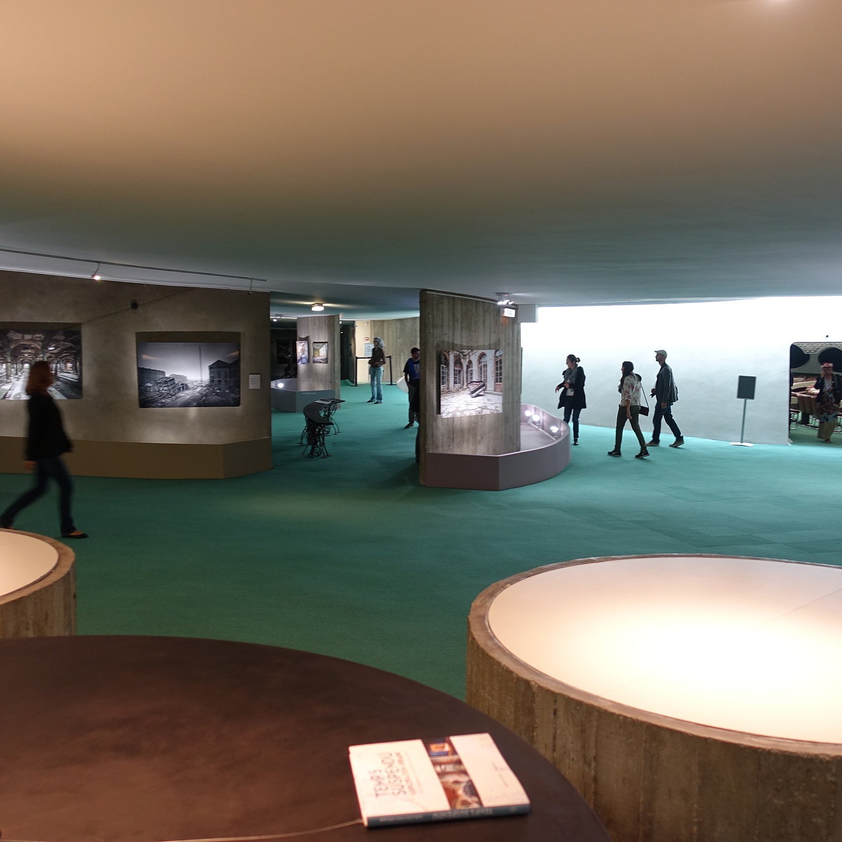 L'image présente un espace d'exposition, probablement dans un musée. On peut y voir des murs de couleur claire avec des photographies accrochées. Le sol est recouvert d'un tapis vert, créant une atmosphère accueillante. Des visiteurs se déplacent dans cet espace, explorant les œuvres exposées. Au premier plan, il y a plusieurs tables ou surfaces circulaires, apparemment utilisées pour des brochures ou d'autres informations. L'éclairage est doux et met en valeur les œuvres sur les murs.