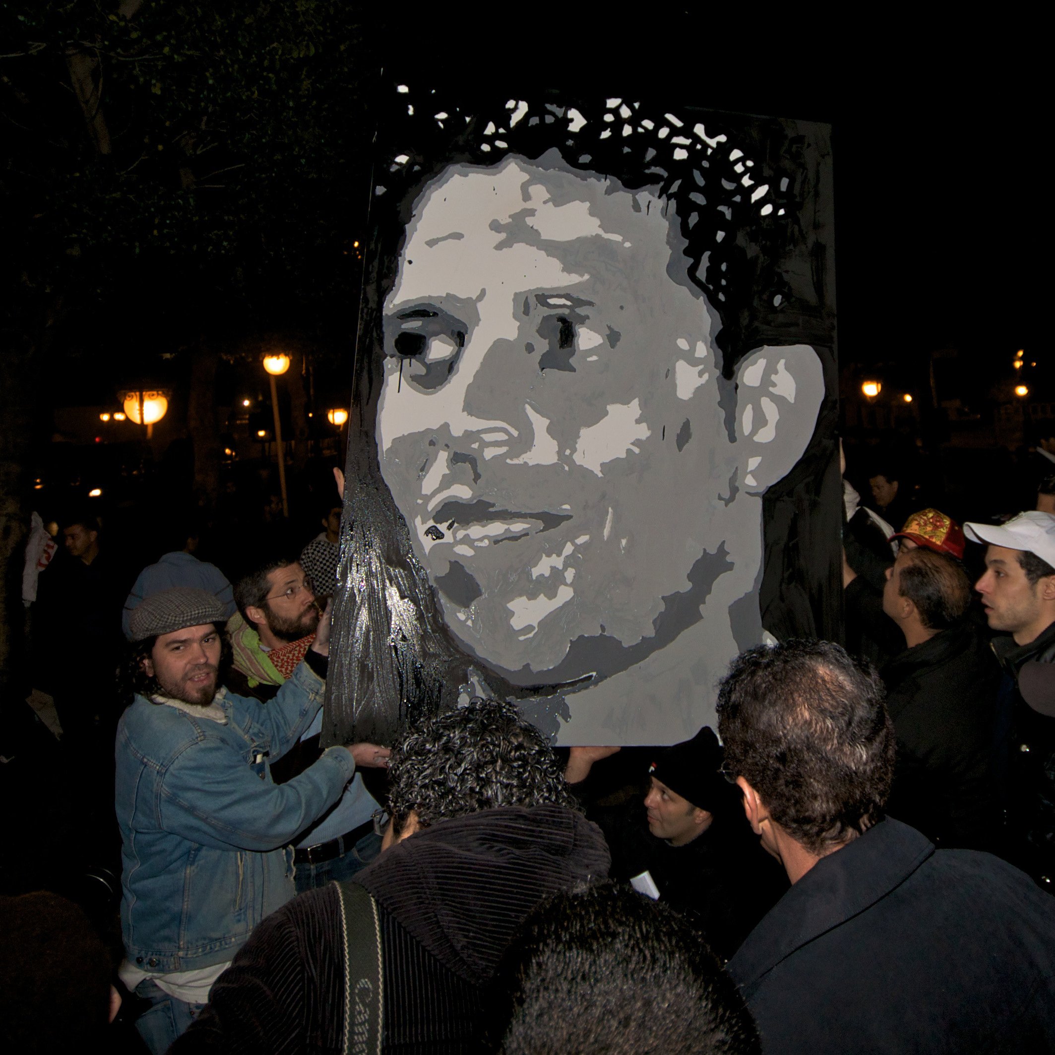 L'image montre un groupe de personnes rassemblées dans un environnement nocturne. Au centre, une grande affiche ou un portrait d'un homme est visible, avec des couleurs sombres et une expression marquante. Les personnes semblent engagées et intéressées par le portrait qu'elles tiennent, ce qui suggère qu'il s'agit d'une manifestation ou d'un hommage. L'ambiance nocturne est accentuée par l'éclairage des lampadaires en arrière-plan.