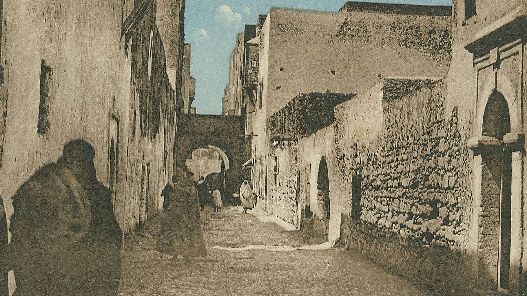 L'image représente une ruelle étroite et pavée, probablement dans une ville historique. Les murs des bâtiments sont en pierre, avec des textures vieilles et usées, suggérant un certain âge. On peut apercevoir des silhouettes de personnes marchant, certaines vêtues de longues robes traditionnelles. À l'arrière-plan, un arc architectural est visible, ajoutant une touche d'authenticité à l'environnement. Le ciel est dégagé, apportant une lumière douce à la scène. L'atmosphère semble paisible et évoque un lieu chargé d'histoire.