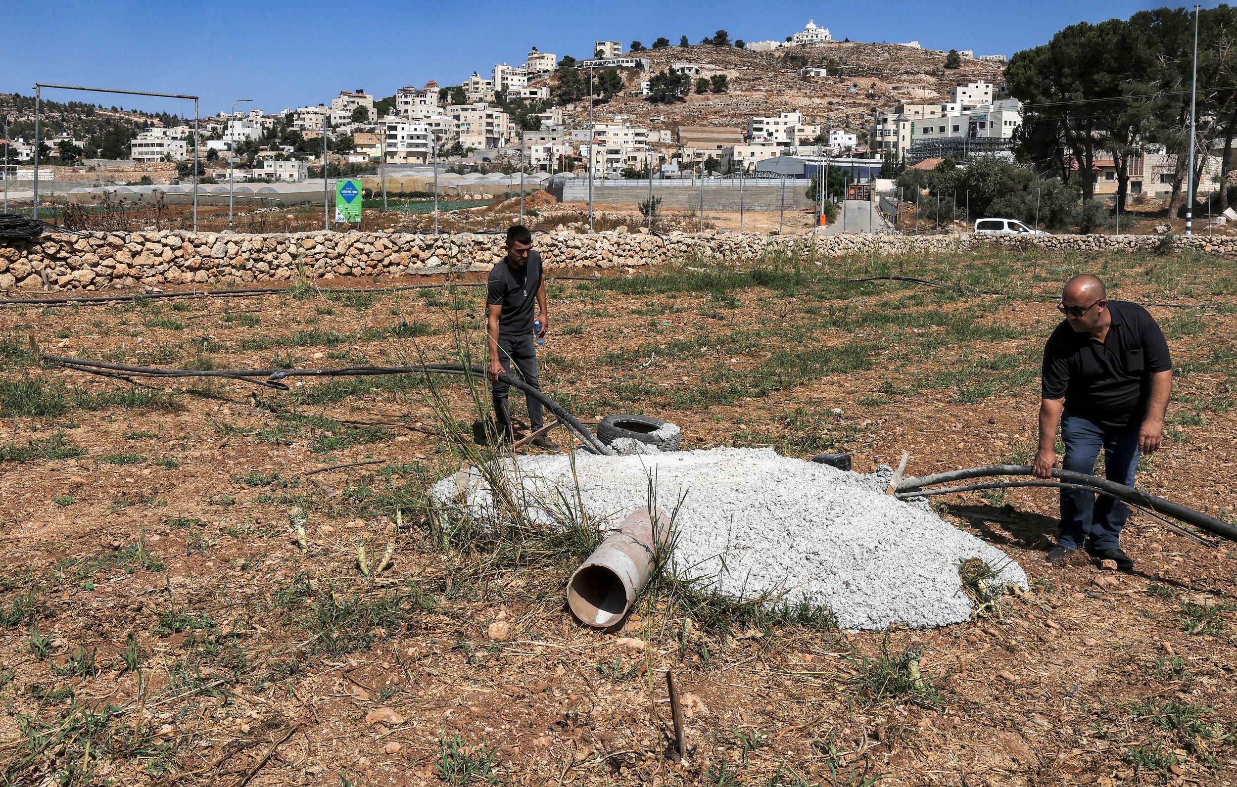 در این تصویر، دو مرد در حال کار بر روی یک زمین کشاورزی هستند. آنها مشغول بررسی یا نصب لوله‌هایی هستند که به یک سازه بتنی متصل شده است. درختان و گیاهان در اطراف وجود دارند و در پس‌زمینه، ساختمانی و تپه‌هایی دیده می‌شوند که حاکی از یک منطقه مسکونی است. آسمان آبی و صاف به نظر می‌رسد.