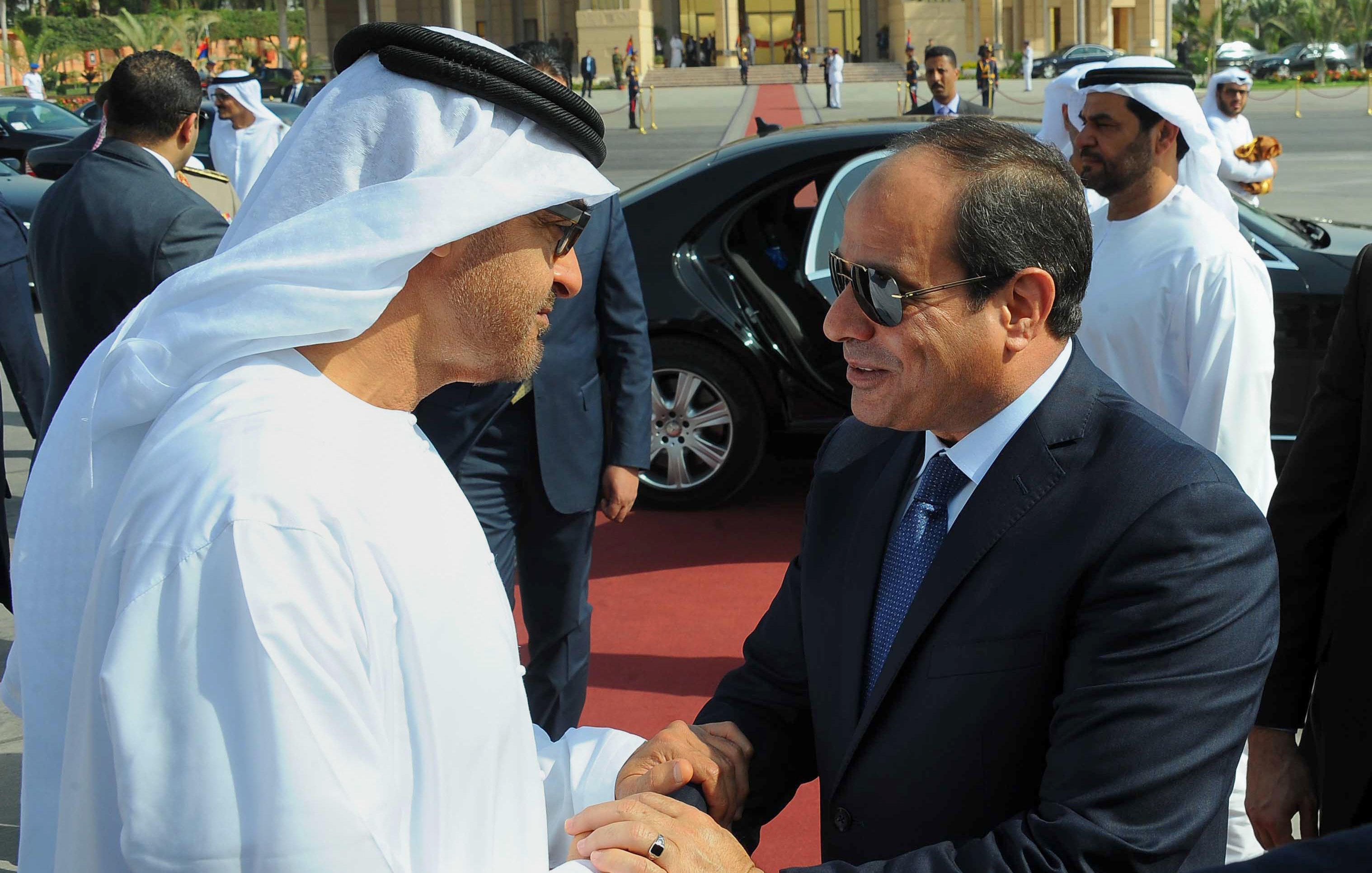 تظهر في الصورة شخصيات يرتديان ملابس تقليدية. الشخص على اليسار يرتدي الثوب الإماراتي الأبيض والغترة، بينما الشخص على اليمين يرتدي بدلة رسمية. يبدو أنهم يتصافحان في مناسبة رسمية، مع وجود سيارات فاخرة في الخلفية ومجموعة من الحراس أو الأشخاص الآخرين. تعكس الصورة أجواء من التعاون والاحترام المتبادل.