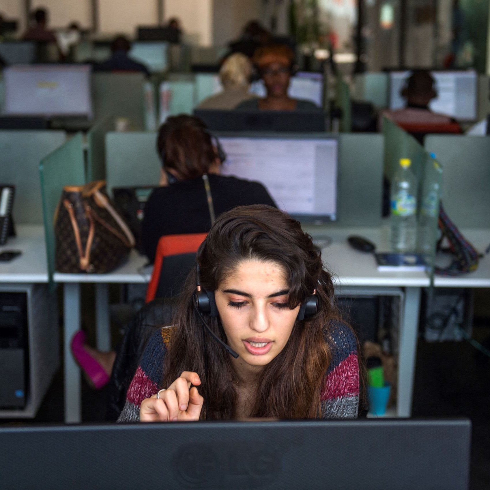 L'image montre un bureau avec plusieurs employés travaillant sur des ordinateurs. Au premier plan, une jeune femme porte un casque et semble concentrée sur son écran. Elle gesticule avec une main, probablement en train de communiquer avec un client. En arrière-plan, d'autres personnes sont visibles, toutes occupées à leurs tâches. L'environnement a l'air moderne, avec des bureaux en open space et des équipements de travail.