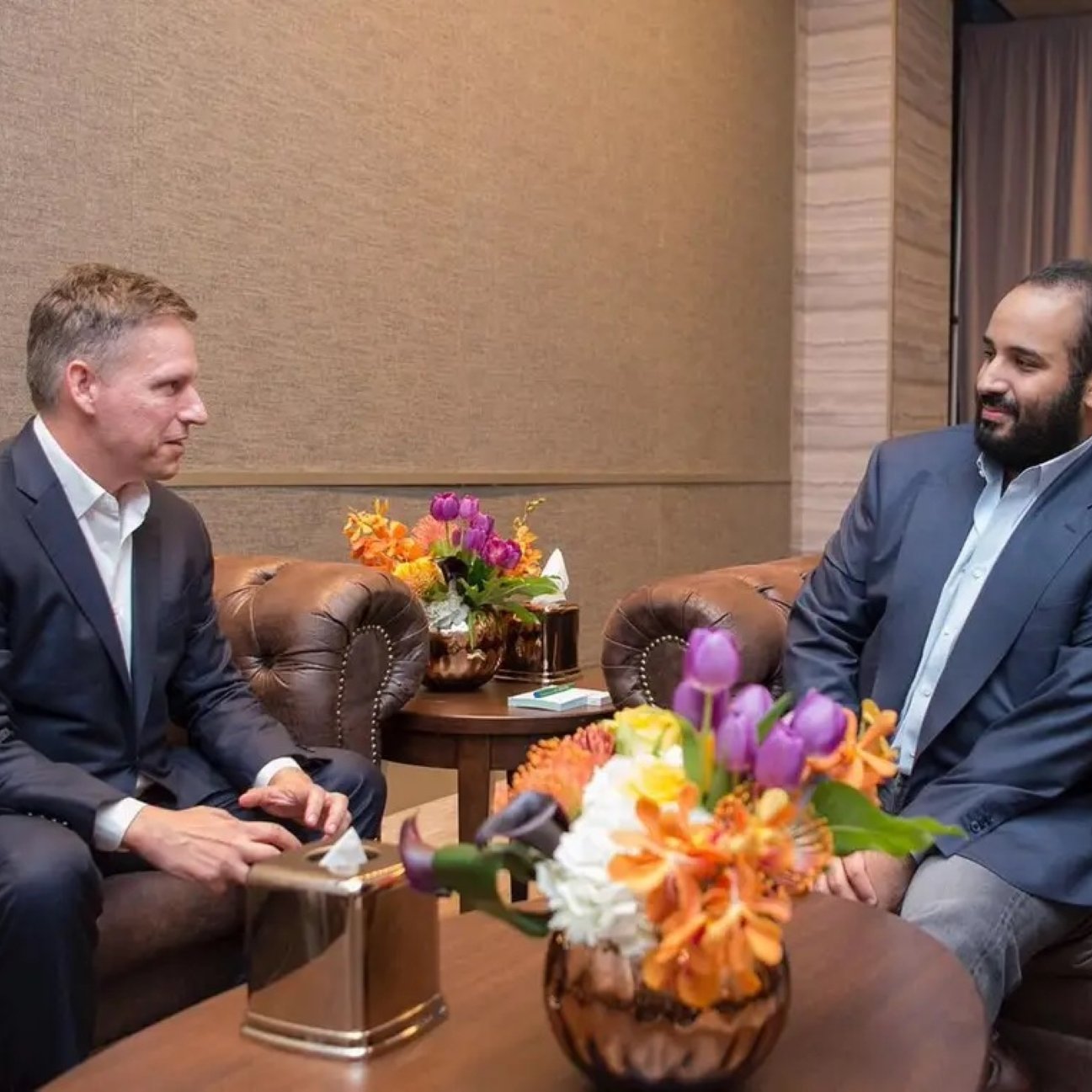 Two men in suits sit in a room, conversing near a floral arrangement on a table.