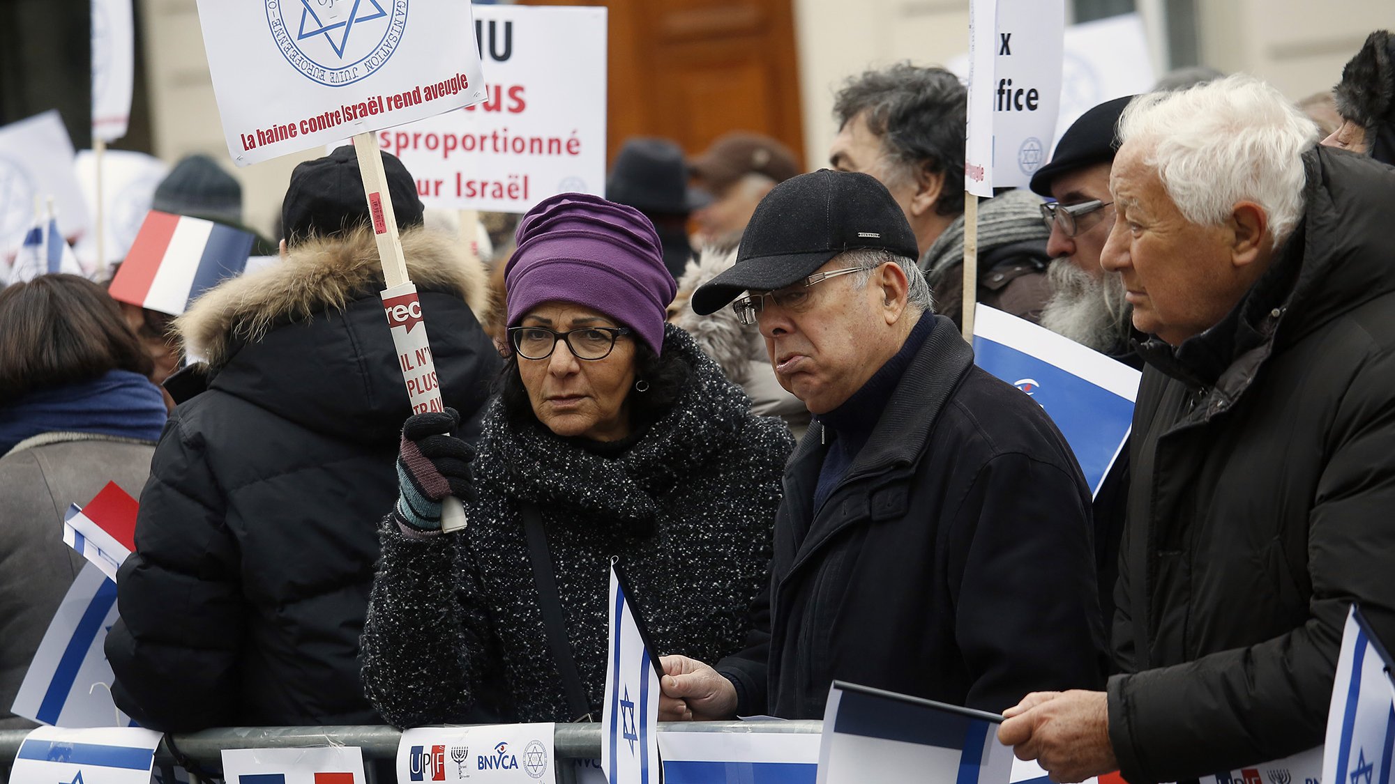 La imagen muestra a un grupo de personas participando en una manifestación. Los manifestantes sostienen pancartas y banderas, muchas de ellas con símbolos relacionados con Israel y Francia. Los asistentes varían en edad y están vestidos de manera variada, algunos con abrigos y gorros, lo que sugiere que es un día frío. El ambiente parece ser de protesta, con mensajes que expresan opiniones sobre el conflicto en Israel.