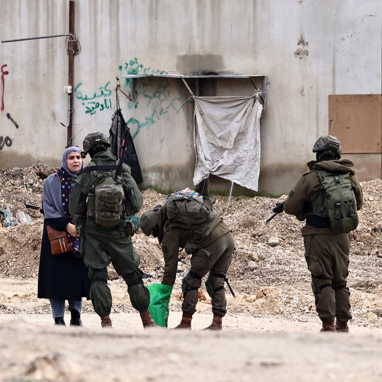 L'image montre une femme vêtue d'une robe sombre, se tenant à côté d'une rue. Devant elle, plusieurs soldats en uniforme sont regroupés, certains sont en train de fouiller un sac, tandis que d'autres observent la femme. En arrière-plan, on peut voir un mur avec des graffitis et des débris sur le sol.