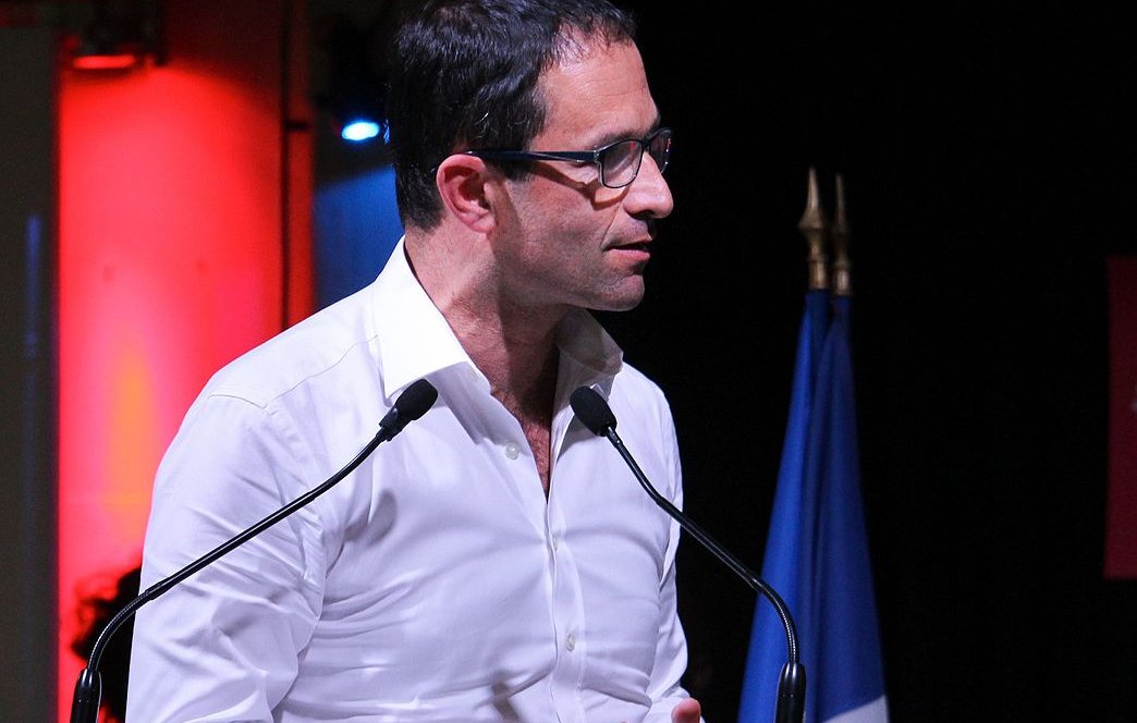 L'image montre un homme debout, parlant à un podium avec le nom "Benoît Hamon 2017" affiché sur le devant. Il porte une chemise blanche et des lunettes, et il semble s'exprimer avec passion. En arrière-plan, on peut voir des éclairages colorés, notamment en rouge, et un drapeau français. L'homme semble engagé dans un discours ou une présentation.