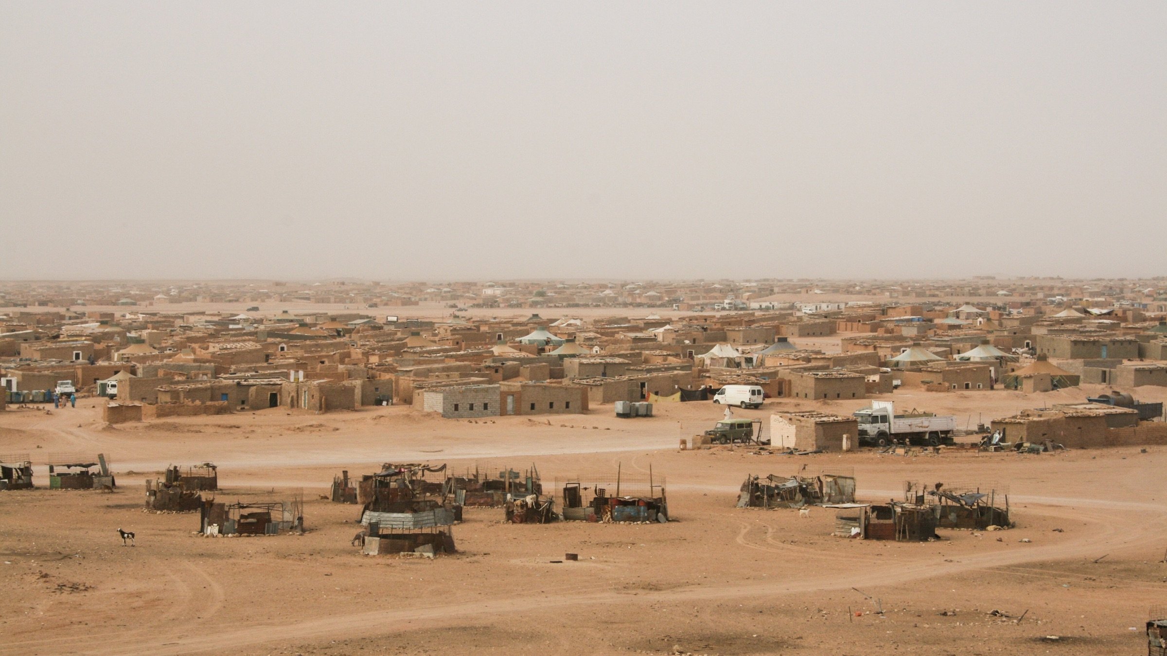 L'image montre un paysage désertique, probablement dans une région aride. On peut voir une vaste étendue de terre sèche, avec des habitations simples construites en terre et en matériaux naturels, disséminées dans le paysage. Certaines constructions semblent être regroupées, tandis que d'autres sont isolées. L'atmosphère est probablement chaude et poussiéreuse, avec un ciel clair ou légèrement embrumé. Les couleurs dominantes sont des tons de beige, de marron et de jaune, typiques des déserts.