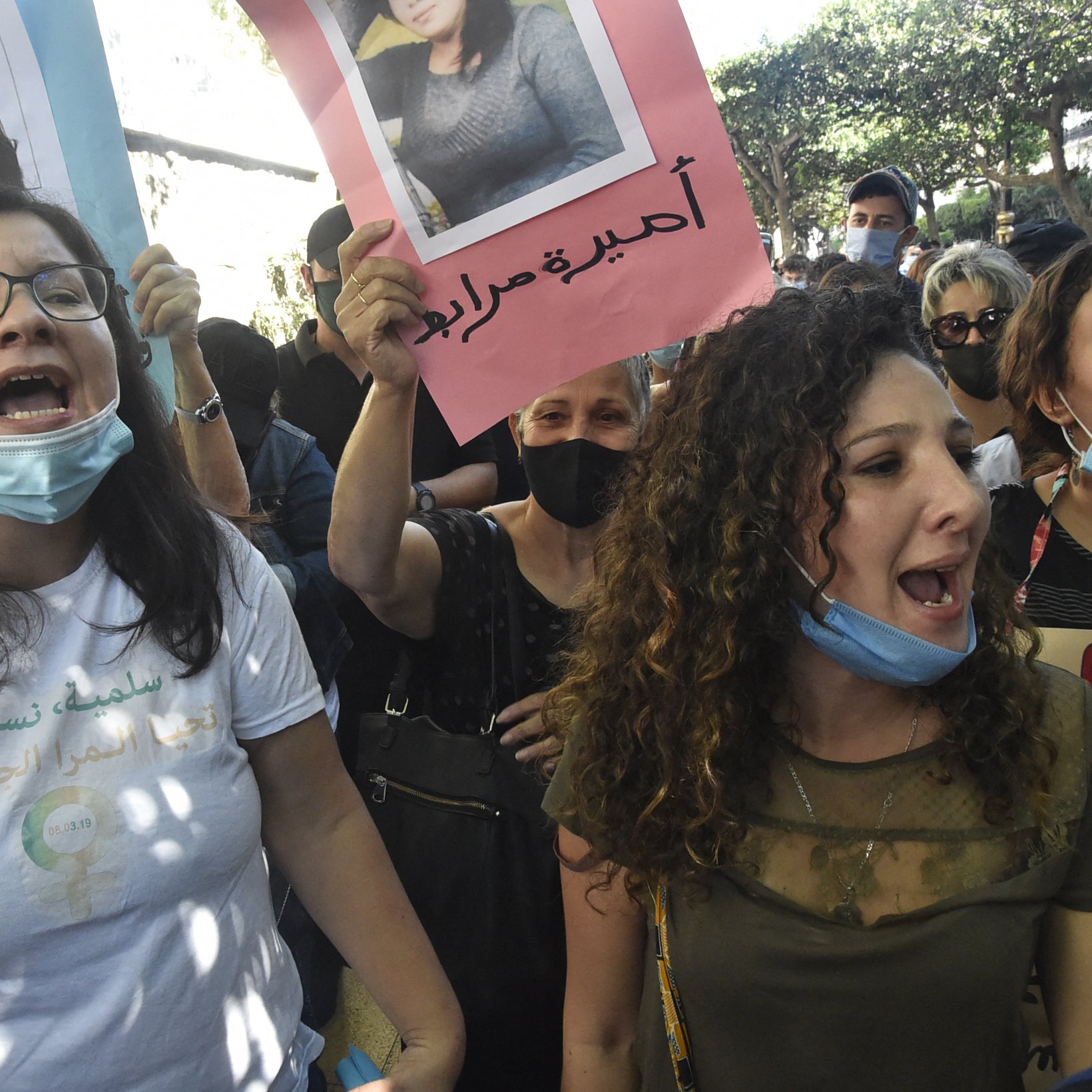 L'immagine mostra un gruppo di persone che partecipa a una manifestazione. Alcuni manifestanti tengono in mano cartelli con fotografie e slogan, mentre gridano in segno di protesta. Le persone indossano mascherine e sembrano essere molto coinvolte emotivamente, esprimendo le loro richieste con forza. Il contesto sembra essere urbano, con alberi e edifici sullo sfondo.