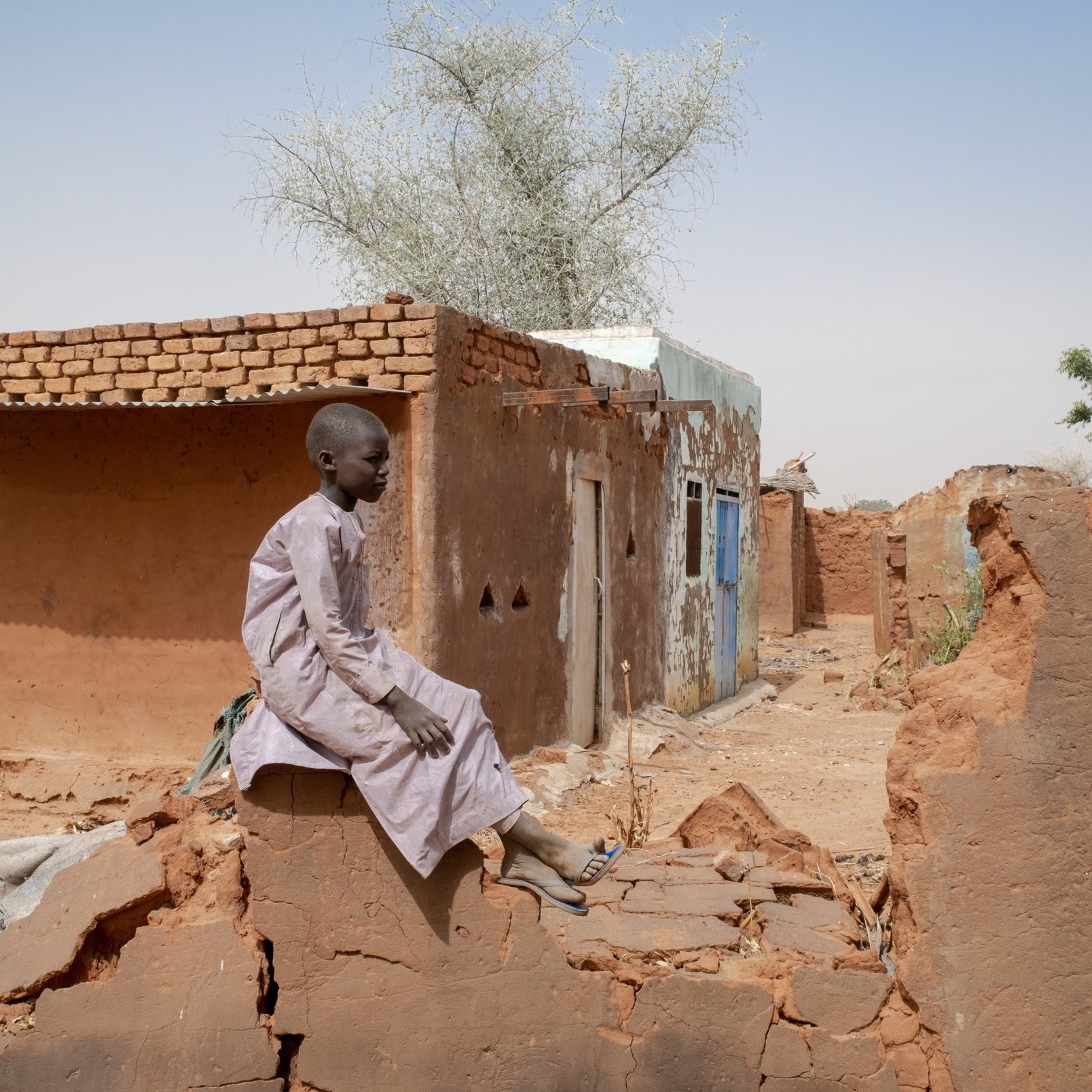 L'image montre un jeune garçon assis sur de décombres dans une région désertique. Il porte une tunique traditionnelle et semble pensif. En arrière-plan, il y a des bâtiments en terre cuite, dont certains sont partiellement détruits. Le paysage est aride, avec quelques arbres éparpillés, et le ciel est clair. L'atmosphère dégage une impression de solitude et de résilience face aux conditions difficiles.