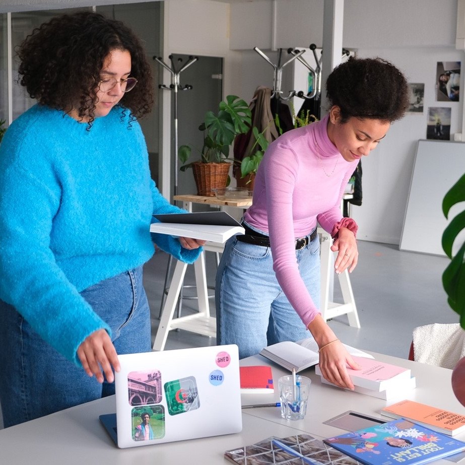 The image shows two women in a modern workspace, engaged in an activity together. One woman, wearing a bright blue sweater, is standing and holding a stack of papers or a folder. The other woman, dressed in a pink top, is bending over a table, arranging or placing items. The table is cluttered with books, a laptop, and other materials, indicating a creative environment. There are plants in the background, adding a touch of greenery to the space. The overall atmosphere appears collaborative and productive.
