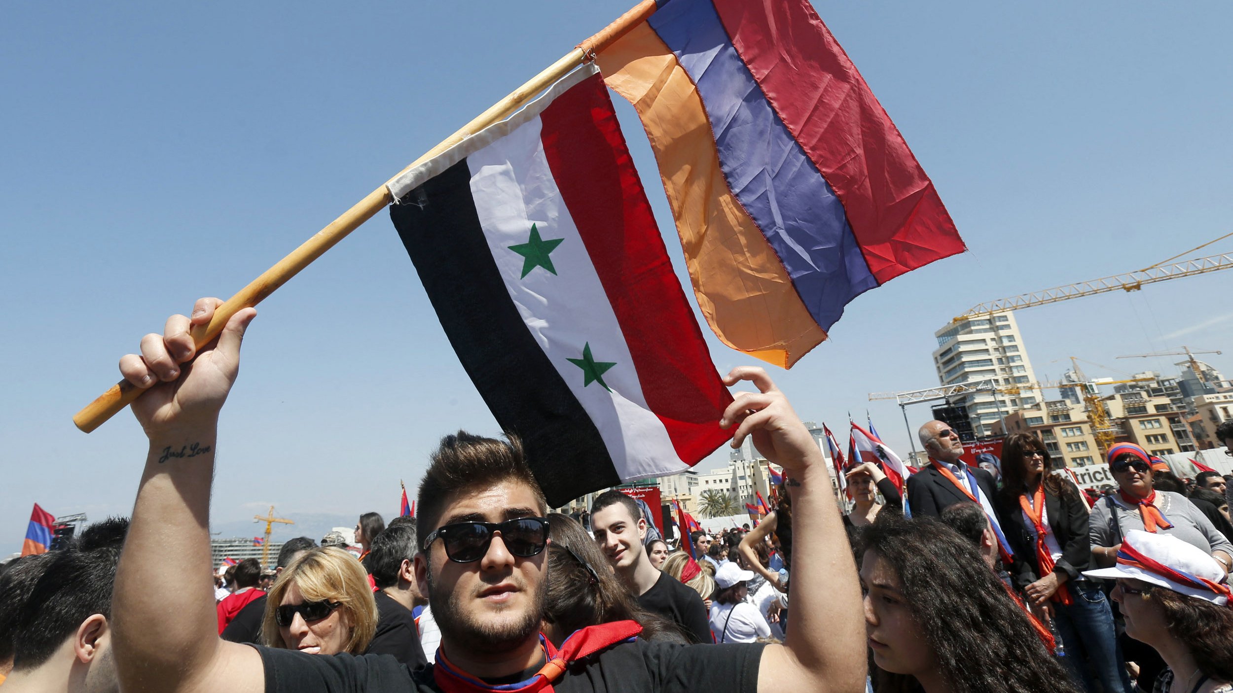 L'image montre un groupe de personnes participant à une manifestation ou un rassemblement. Un homme au premier plan tient fièrement trois drapeaux : le drapeau syrien, un drapeau avec trois étoiles vertes sur fond noir et rouge, ainsi qu'un drapeau arménien. L'ambiance semble festive et engagée, avec de nombreuses personnes en arrière-plan, probablement partageant des sentiments communs ou soutenant une cause. Le ciel est clair, ce qui ajoute à l'énergie de l'événement.