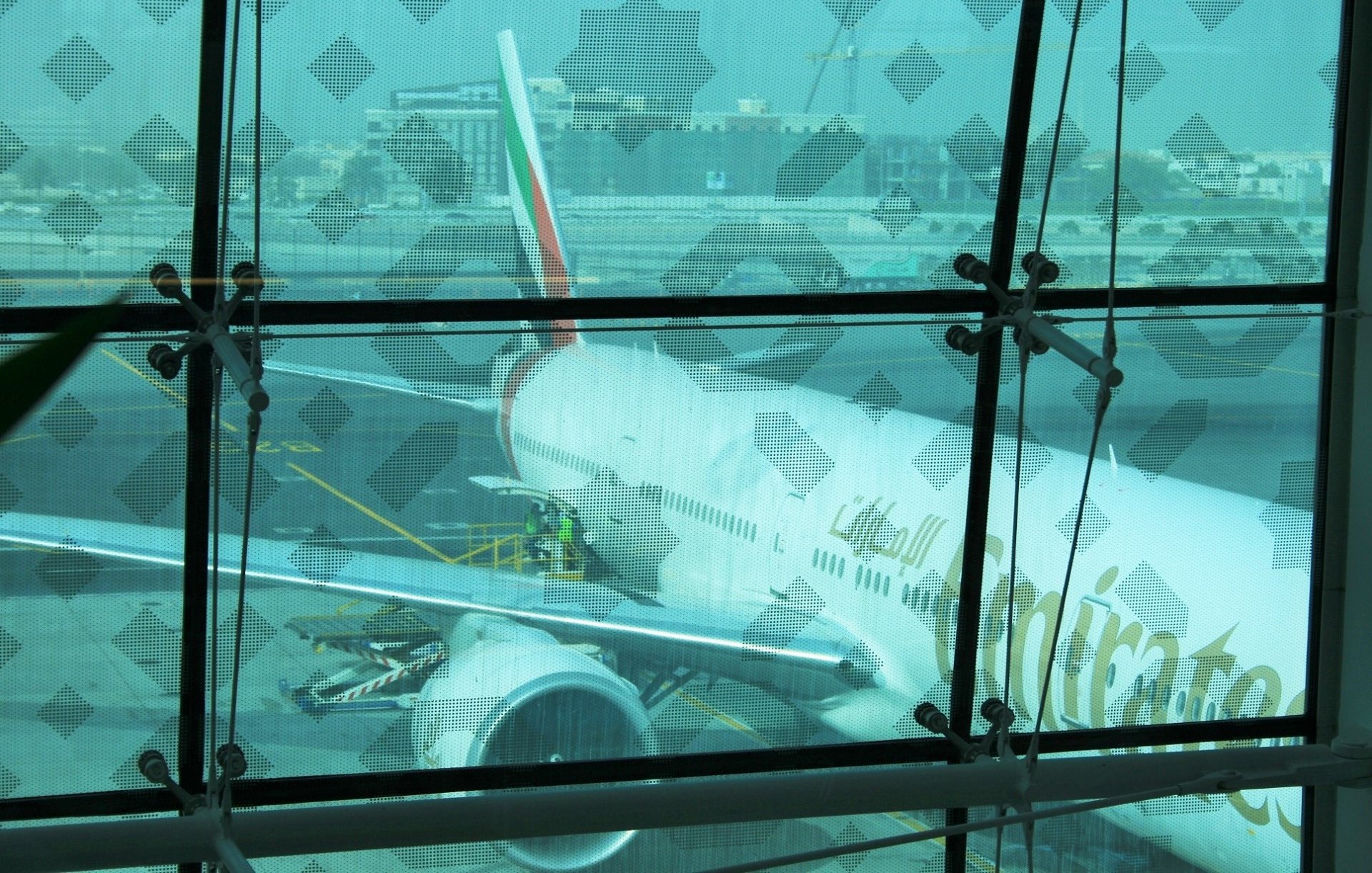 The image shows an airplane belonging to Emirates Airlines parked at an airport. The aircraft appears to be a large model, likely a Boeing 777, as indicated by the shape of the fuselage and engines. You can see maintenance personnel working on the plane, and the background features airport buildings and infrastructure. The scene is framed by a glass wall, which has a patterned overlay, adding an artistic element to the view. The overall atmosphere seems busy and operational, typical for a major airport.