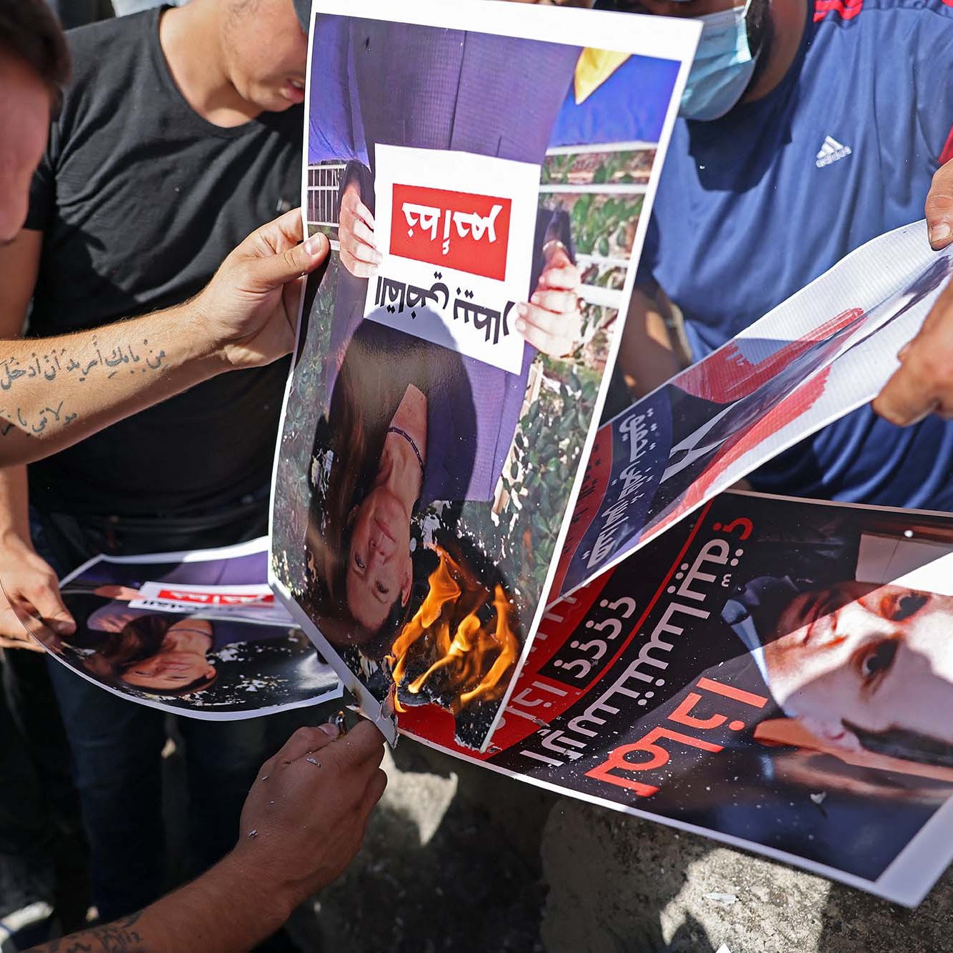 Nell'immagine si vedono diverse persone che stanno bruciando dei manifesti. I manifesti ritraggono volti, probabilmente di una figura pubblica, e sono esposti a una fiamma. Le persone sembrano partecipare attivamente a questa protesta o atto simbolico, esprimendo il loro discontento. L'atmosfera è di intensa emozione e opposizione.