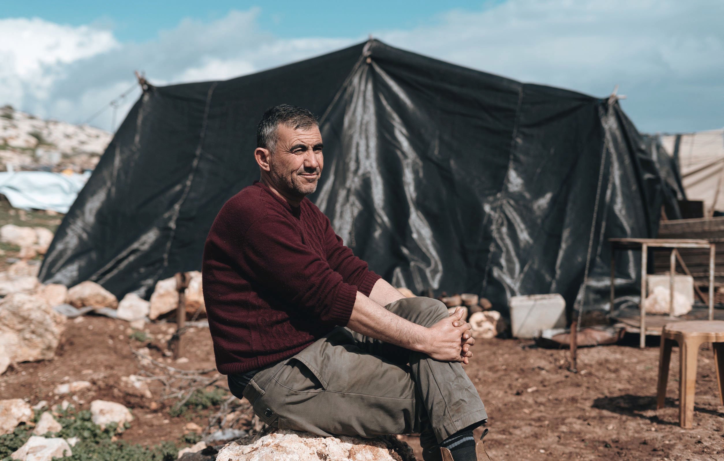 L'image montre un homme assis sur une pierre, avec un air détendu, près d'une tente noire. Il porte un pull rouge et des pantalons kaki. En arrière-plan, on peut apercevoir des tentes et des rochers dans un environnement naturel. Le ciel est partiellement nuageux. L'atmosphère semble calme, malgré les conditions de vie évidentes.