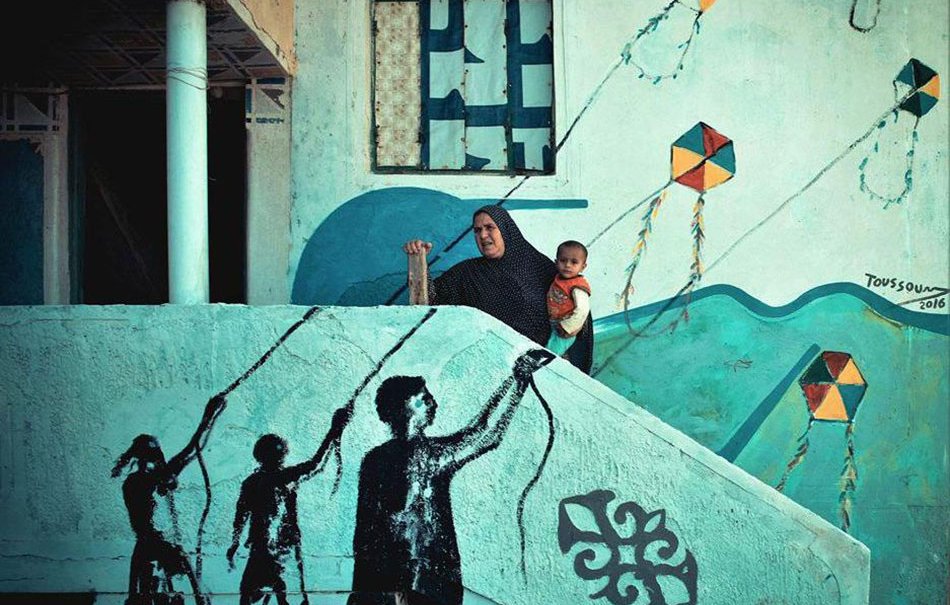 L'image montre une femme, vêtue d'un hijab, tenant un enfant dans ses bras, debout sur une marche d'escalier. En arrière-plan, on peut voir un mur peint avec des motifs colorés et des cerfs-volants. Sur la marche, il y a une fresque représentant des silhouettes de personnes tenant des cerfs-volants, créant un contraste entre l'art mural et la scène réelle. L'ensemble de la composition évoque un sentiment de nostalgie et de connexion entre le passé et le présent.