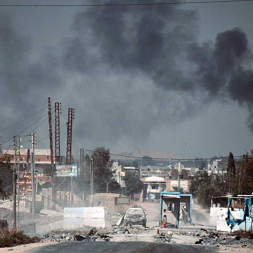 L'image montre une scène de destruction et de conflit. On y voit une route endommagée, avec des débris et des voitures laissées à l'abandon. À l'arrière-plan, un épais nuage de fumée noire s'élève, probablement en raison d'incendies ou d'explosions. Le paysage semble désolé, avec des bâtiments en ruine et une atmosphère de désolation. La présence de poteaux électriques et de panneaux publicitaires ajoute au contexte urbain de l'environnement. L'ensemble de l'image évoque des situations de crise et de guerre.
