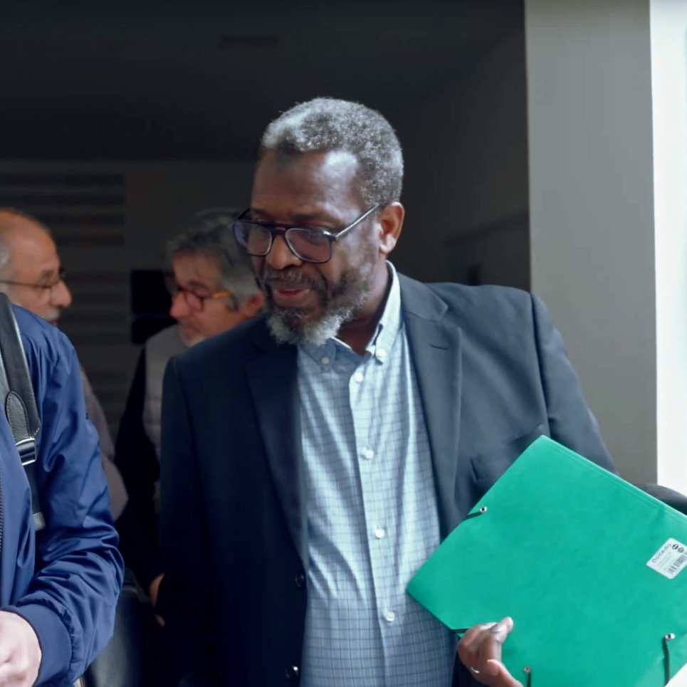 The image shows two men engaged in conversation as they exit a building. One man, wearing glasses and a navy jacket, is looking at his phone while the other, dressed in a black suit, is holding a green folder. In the background, there are other people visible, indicating a busy environment, possibly a court or conference setting. The signage indicates different audience rooms, adding context to the location.