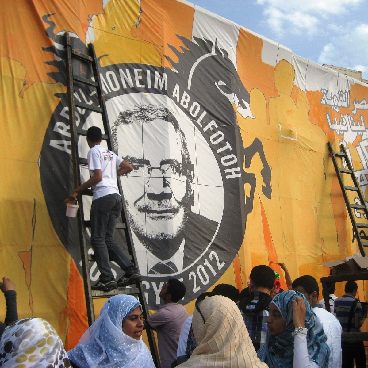 تظهر الصورة مجموعة من الأشخاص يعملون على رسم وإعداد لوحة ضخمة تحمل صورة وشعار أحد المرشحين الانتخابيين، حيث يقوم بعضهم بتلوين اللوحة باستخدام سلالم. الخلفية برتقالية مع تفاصيل أخرى، ويبدو أن الحدث مرتبط بالانتخابات في عام 2012. يظهر أفراد يرتدون ملابس متنوعة، بما في ذلك بعض الأشخاص الذين يرتدون أغطية رأس تحمل ألوان مختلفة. أجواء العمل والاجتهاد تسيطر على المشهد.