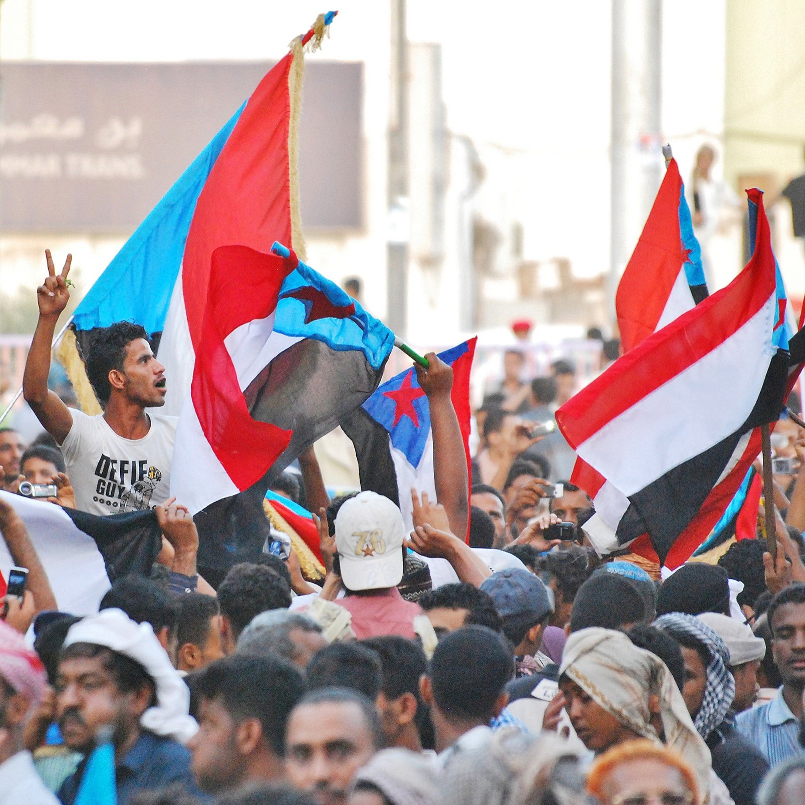 تظهر الصورة حشوداً من المتظاهرين يرفعون أعلاماً ملونة، بما في ذلك الأعلام الحمراء والزرقاء. يبدو أن هناك شعوراً بالحماس والنشاط بين المتظاهرين، حيث يُظهر البعض تحية النصر. يمكن رؤية مجموعة متنوعة من الأشخاص، مما يشير إلى تنوع المشاركين في هذا التجمع. تعكس الصورة أجواءً من التعبير عن الرأي والمشاركة السياسية.