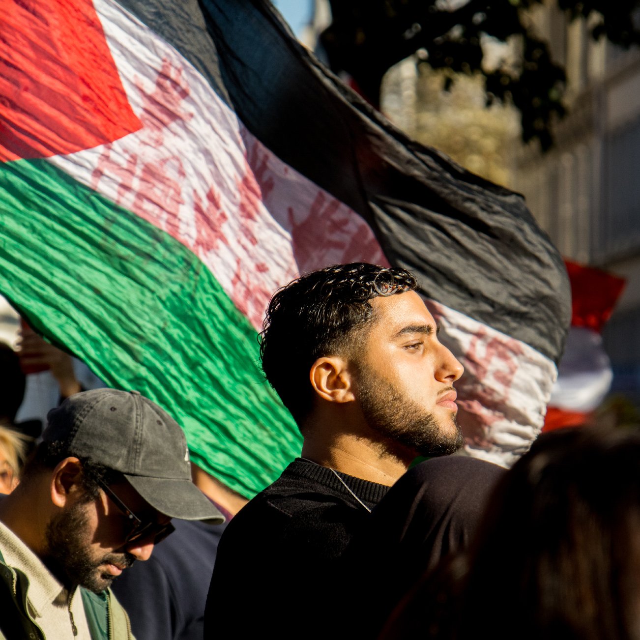L'image montre une foule lors d'une manifestation. On peut voir une personne au premier plan tenant un grand drapeau aux couleurs du pays. Il y a également d'autres manifestants en arrière-plan, dont certains portent des lunettes de soleil et d'autres ont des expressions engagées. La lumière du soleil semble créer une ambiance dynamique et engagée, accentuant l'émotion de l'événement.