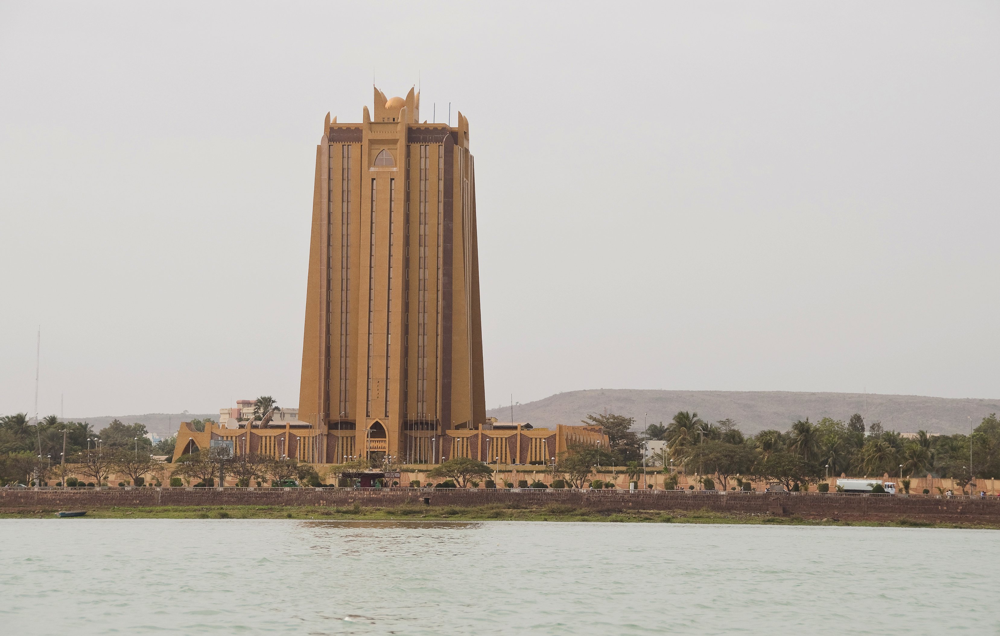 این تصویر نمای یک ساختمان بلند و برجسته را نشان می‌دهد که به رنگ زرد مایل به قهوه‌ای است. ساختمان دارای طراحی خاص و مدرنی است و با چندین طبقه و نوک‌های تیز در بالای آن مشخص می‌شود. در پیش‌زمینه، آب وجود دارد که به زیبایی تصویر می‌افزاید، و در پس‌زمینه، تپه‌ها و نواحی سبز دیده می‌شود. فضای عمومی اطراف ساختمان نیز مشخص است.