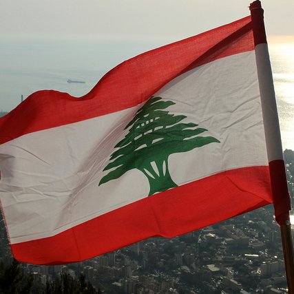 L'image montre le drapeau du Liban flottant au vent, avec un fond qui semble représenter une vue panoramique d'une ville côtière. Le drapeau se compose de trois bandes horizontales : rouge en haut et en bas, et une bande blanche au centre avec un grand cèdre vert, emblème national du Liban. L'horizon présente une mer scintillante, probablement au coucher du soleil, ce qui ajoute une ambiance paisible et naturelle à la scène.
