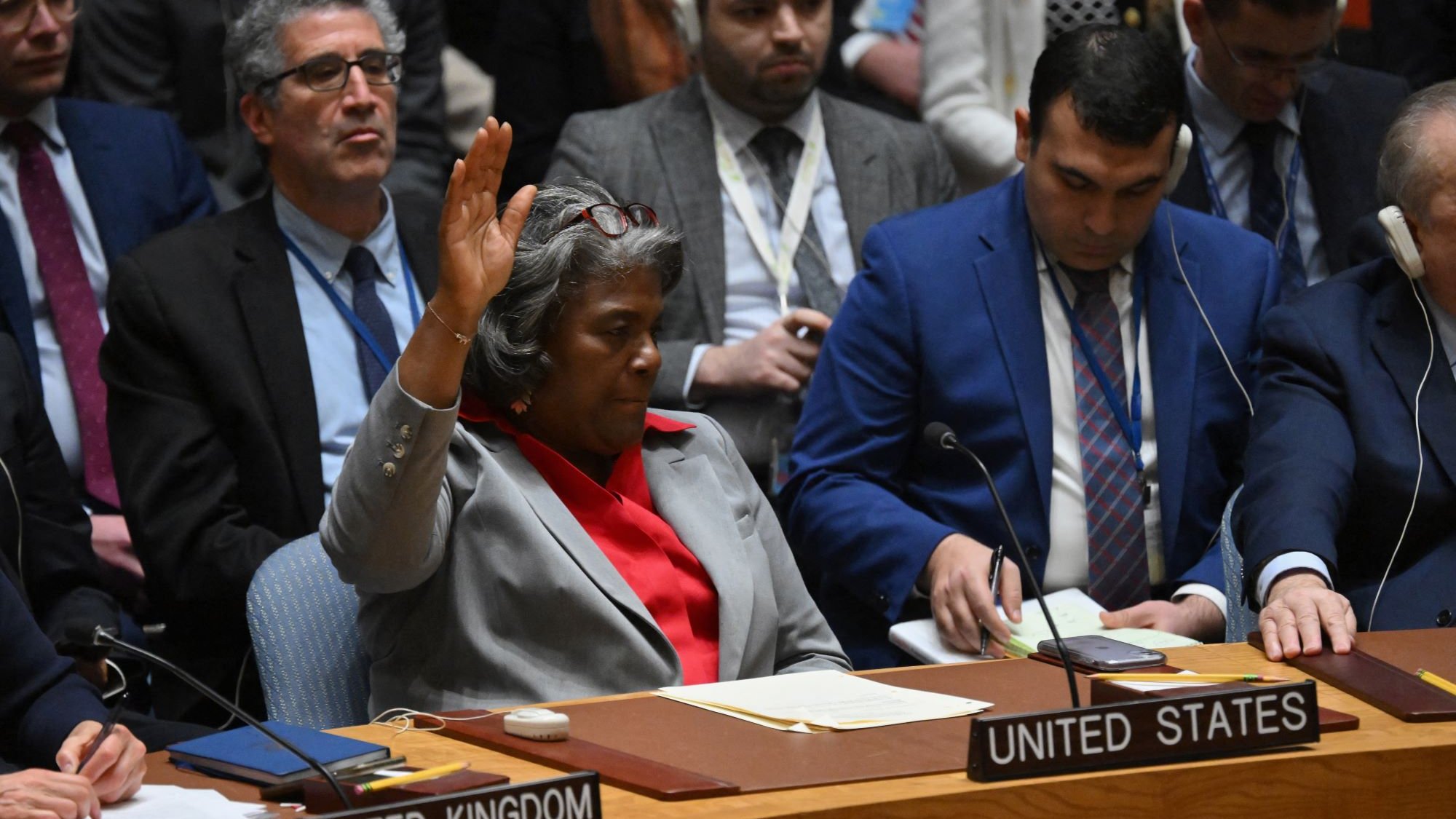 L'immagine mostra un incontro del Consiglio di Sicurezza delle Nazioni Unite. Una donna, che sta al centro della scena, alza la mano, apparentemente per esprimere un'opinione o richiedere la parola. Indossa un blazer grigio con una camicia rossa e sembra essere in fase di comunicazione. Attorno a lei, ci sono diversi rappresentanti di altri paesi, tra cui stati come Regno Unito e altri, seduti con atteggiamenti attenti. L'atmosfera sembra seria e formale, tipica di una riunione diplomatica.