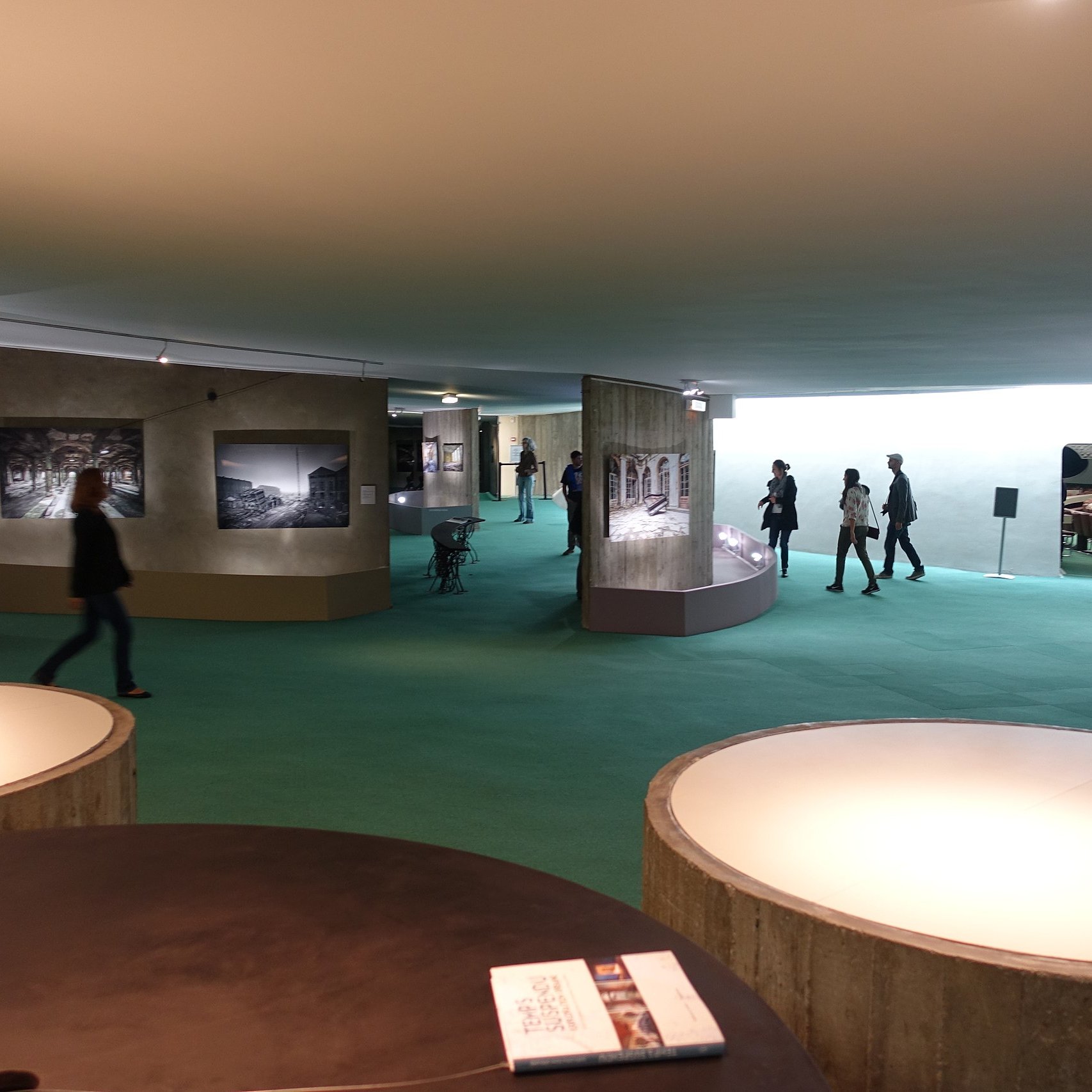The image depicts an interior space, likely an art gallery or museum. The floor is covered with green carpeting, and the walls display a series of black-and-white photographs. Several people are walking through the space, engaged with the exhibits. There are curved display structures that showcase the photographs. The lighting appears bright and modern, creating an inviting atmosphere.