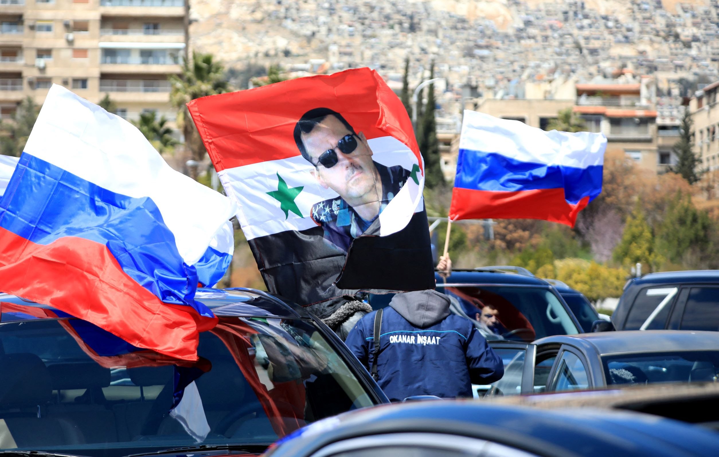 L'image montre une scène dans laquelle des personnes portent des drapeaux russes et un drapeau syrien. Au centre, on peut voir un grand drapeau sur lequel est imprimée une photo d'un homme. La scène semble se dérouler dans une ville, probablement en Syrie, avec des bâtiments en arrière-plan sur une colline. L'atmosphère suggère un rassemblement ou une manifestation, possiblement en soutien à une cause politique.