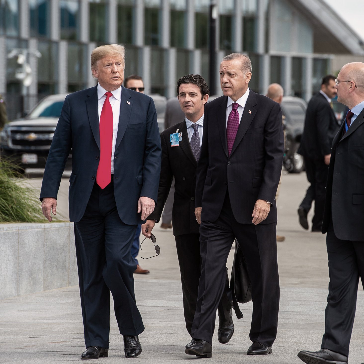 L'image montre un groupe d'hommes en costumes, apparemment dans un cadre formel, probablement lors d'un événement diplomatique. Deux hommes, respectivement en costume noir et rouge, marchent côte à côte, tandis que d'autres les suivent. L'arrière-plan indique un environnement urbain moderne, avec des véhicules et des bâtiments. L'atmosphère semble sérieuse, éventuellement liée à des discussions ou des négociations importantes.