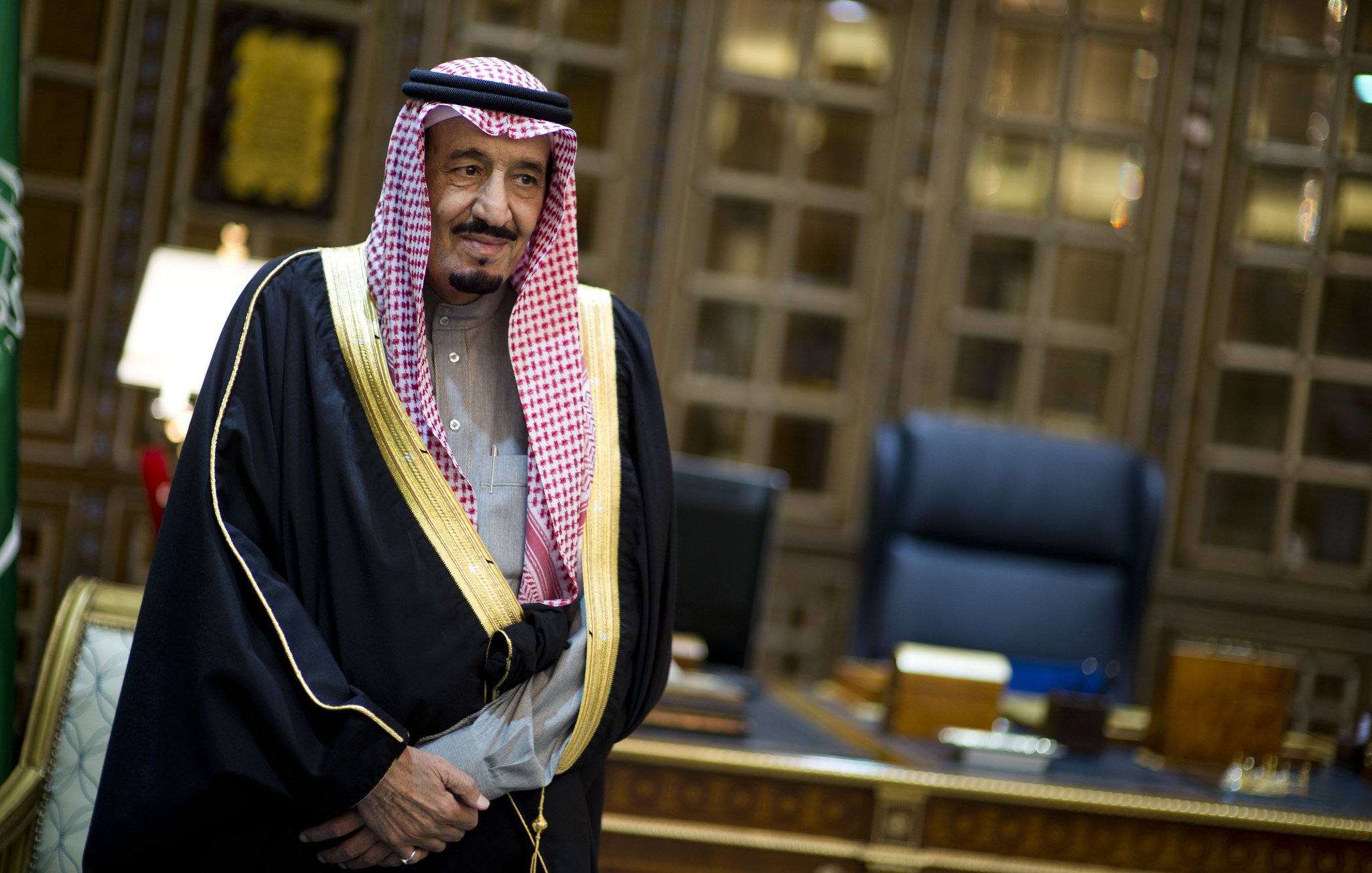 L'image montre un homme en tenue traditionnelle saoudienne, portant une robe noire ornée d'or et un keffieh, une coiffe typique. Il se tient dans un bureau richement décoré, avec des meubles en bois et un éclairage sophistiqué. L'ambiance est formelle et élégante.