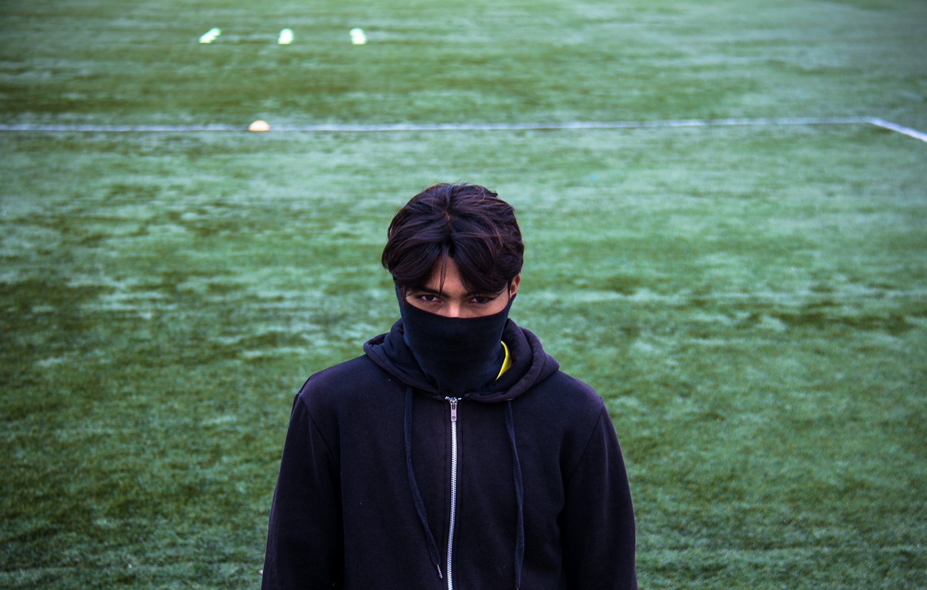 The image features a young person standing on a green sports field, possibly a soccer pitch. They are wearing a black hoodie and have a black mask covering the lower part of their face. The focus is on their intense gaze, and they appear to be looking directly at the camera. The field is marked with white lines and has some yellow cones in the background, suggesting a training or practice session. The overall mood is somewhat mysterious and serious.