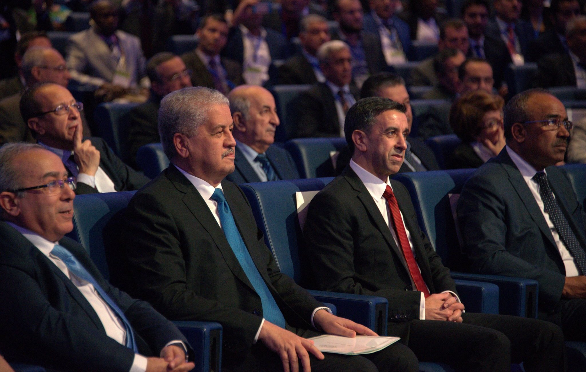 L'image montre un groupe d'hommes assis dans une salle de conférence, apparemment lors d'un événement officiel. Ils portent des costumes sombres, avec une attention particulière sur deux hommes au premier plan, l'un en costume noir avec une cravate bleue, et l'autre en costume noir avec une cravate rouge. En arrière-plan, on peut voir d'autres personnes, probablement des participants ou des invités, attentifs à ce qui se passe sur scène. L'atmosphère semble formelle et sérieuse.