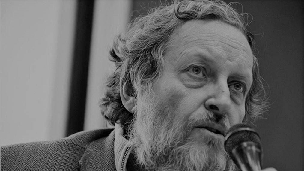 L'immagine mostra un uomo con un aspetto intellettuale. Ha una barba e capelli ricci, e sembra in fase di parlare al microfono. Indossa una giacca grigia e un maglione, trasmettendo un'aria di serietà e riflessione. Lo sfondo è sfocato, mettendo in evidenza il suo volto e la sua espressione concentrata.