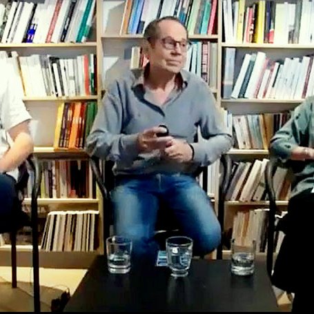 Cette image montre trois personnes assises sur des chaises dans un environnement intérieur, probablement une librairie ou un espace de discussion. À l'arrière-plan, il y a des étagères remplies de livres. La personne à gauche porte une chemise blanche, celle du centre est en chemise grise et celle à droite porte un haut vert. Ils semblent être en train de discuter ou de débattre, avec des verres d'eau devant eux. L'ambiance semble détendue et propice à l'échange d'idées.