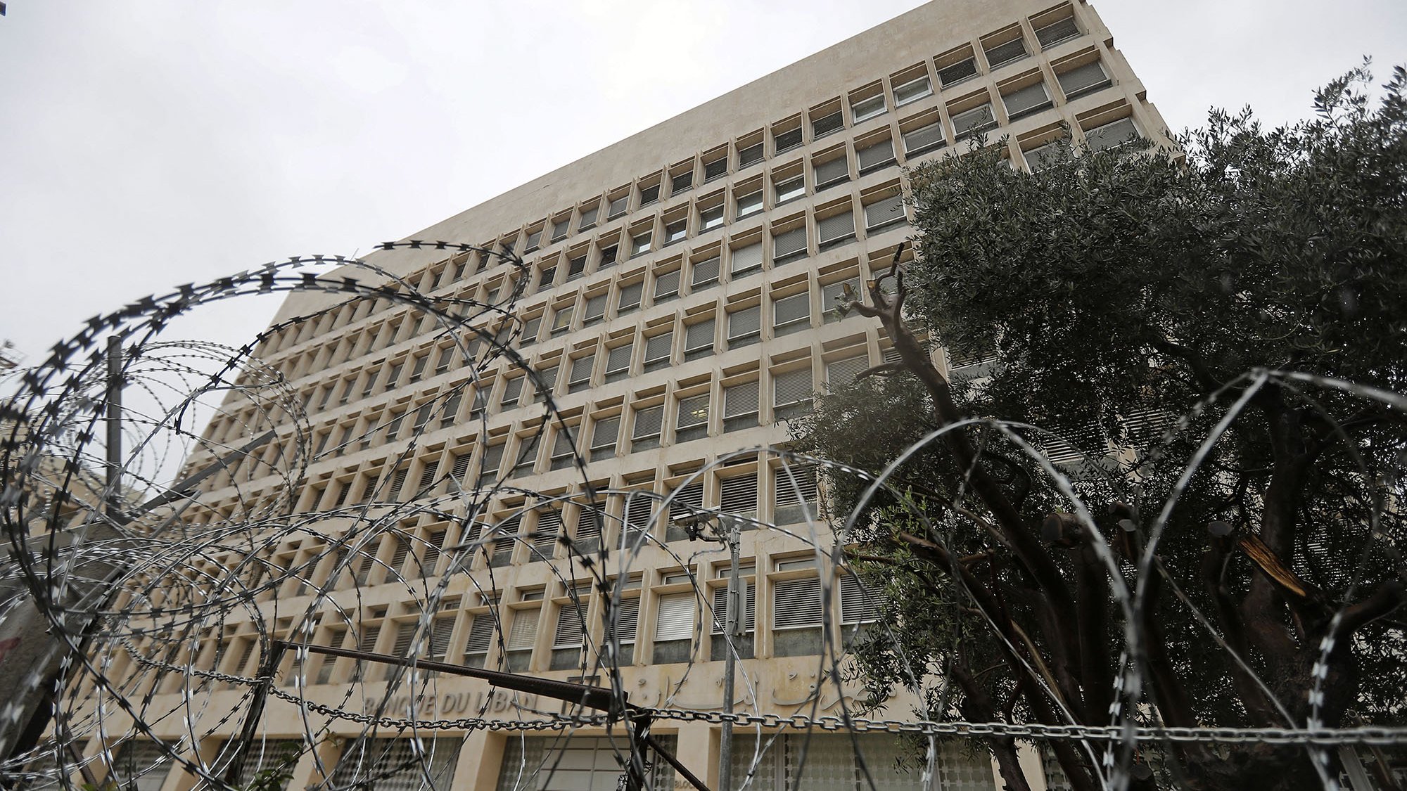 الصورة تظهر مبنى عالٍ يظهر بشكل معماري حديث، ويتواجد في المقدمة سياج من الأسلاك الشائكة. السماء فوق المبنى ملبدة بالغيوم، مما يوحي بجو قاتم. تحيط بالمبنى بعض الأشجار، مما يضيف لمسة خضراء في المشهد. تعكس الصورة شعورًا بالأمان أو القيود، نظراً للسياج الذي يحيط بالمكان.