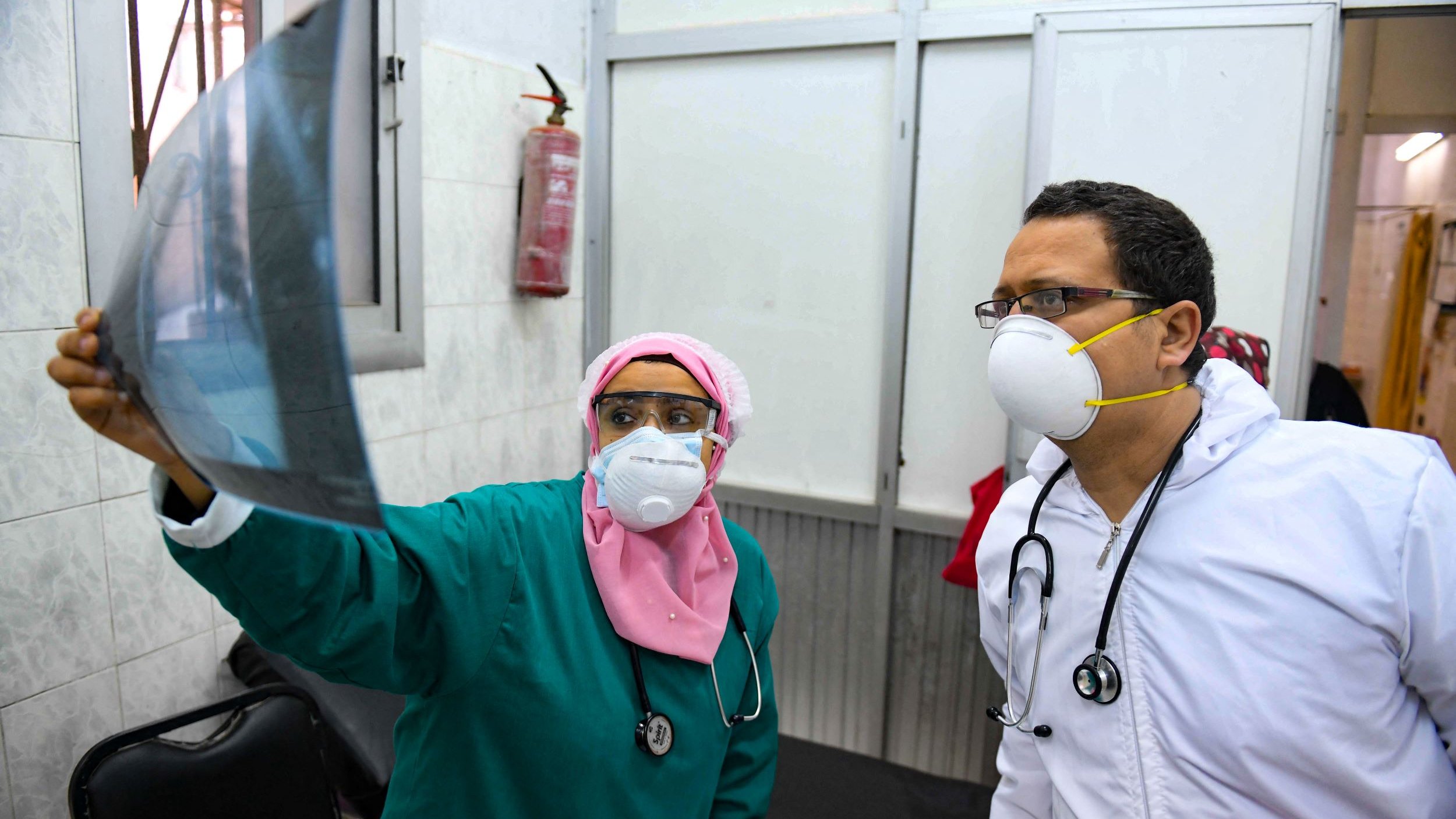 L'image montre deux professionnels de la santé examinant une radiographie. La femme porte un uniforme vert, un masque et un foulard rose, tandis que l'homme est vêtu d'une blouse blanche et porte également un masque. Ils se tiennent dans une pièce médicale, entourés de murs en carrelage. On peut voir un extincteur accroché au mur. L'expression de concentration indique qu'ils discutent sérieusement des résultats de l'examen.