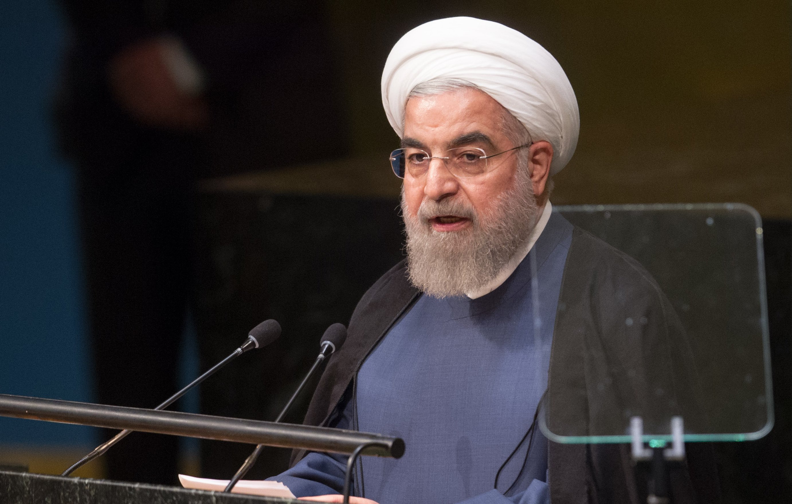 L'image montre un homme parlant à un podium lors d'un événement officiel, probablement à l'ONU. Il porte un costume sombre avec un turban blanc et une barbe. L'homme semble s'adresser à un public, et il y a des microphones devant lui. L'environnement est formel, avec un fond flou qui pourrait représenter un auditorium ou une salle de conférence.
