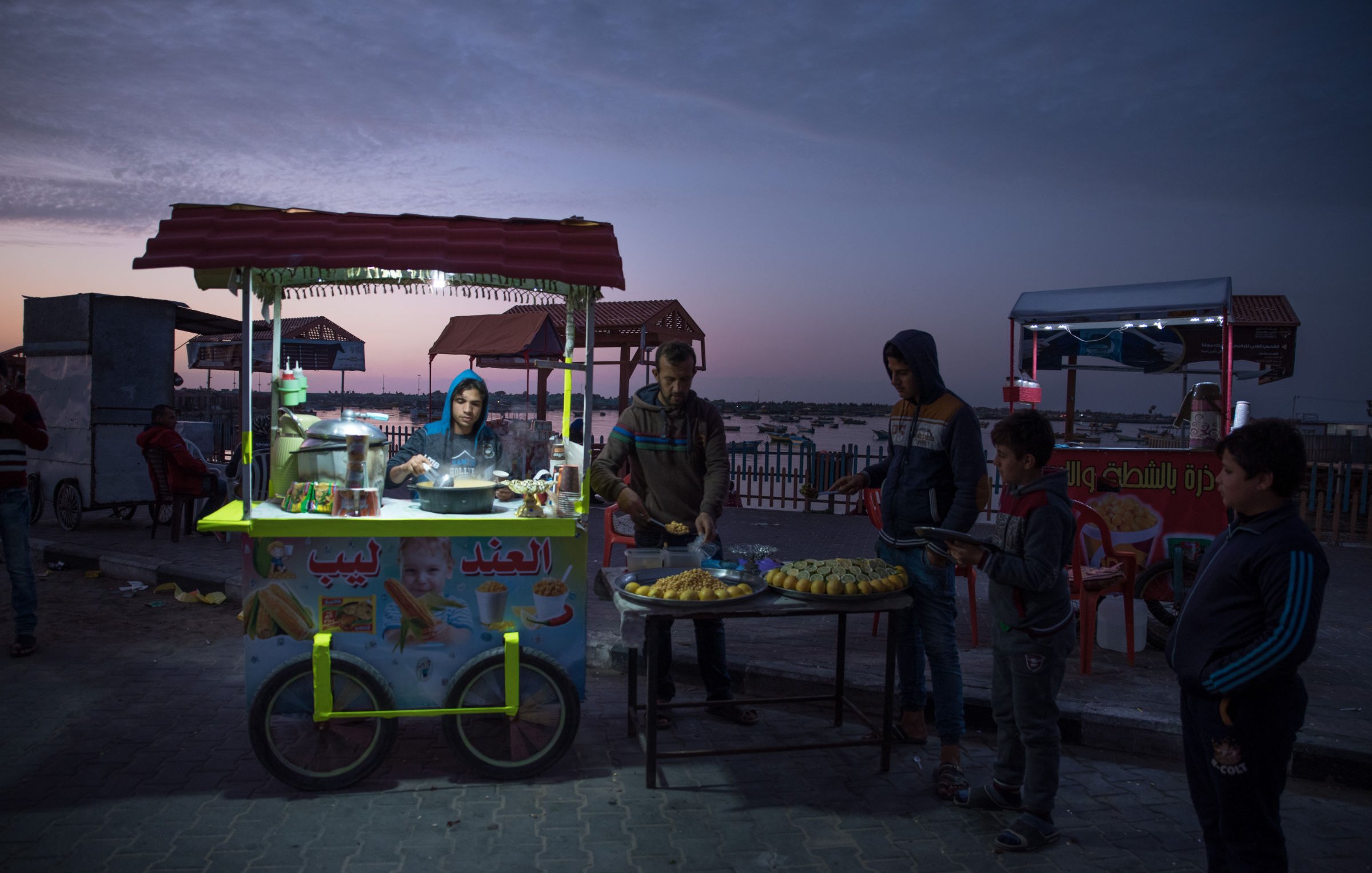 L'image montre un stand de nourriture de rue à la tombée de la nuit, avec un ciel coloré par le crépuscule. Au premier plan, un vendeur prépare des aliments, tandis que plusieurs jeunes clients attendent. Le stand est décoré avec des affiches et contient des produits alimentaires, tels que des pâtisseries. En arrière-plan, on peut apercevoir d'autres stands illuminés et des chaises, créant une ambiance conviviale.