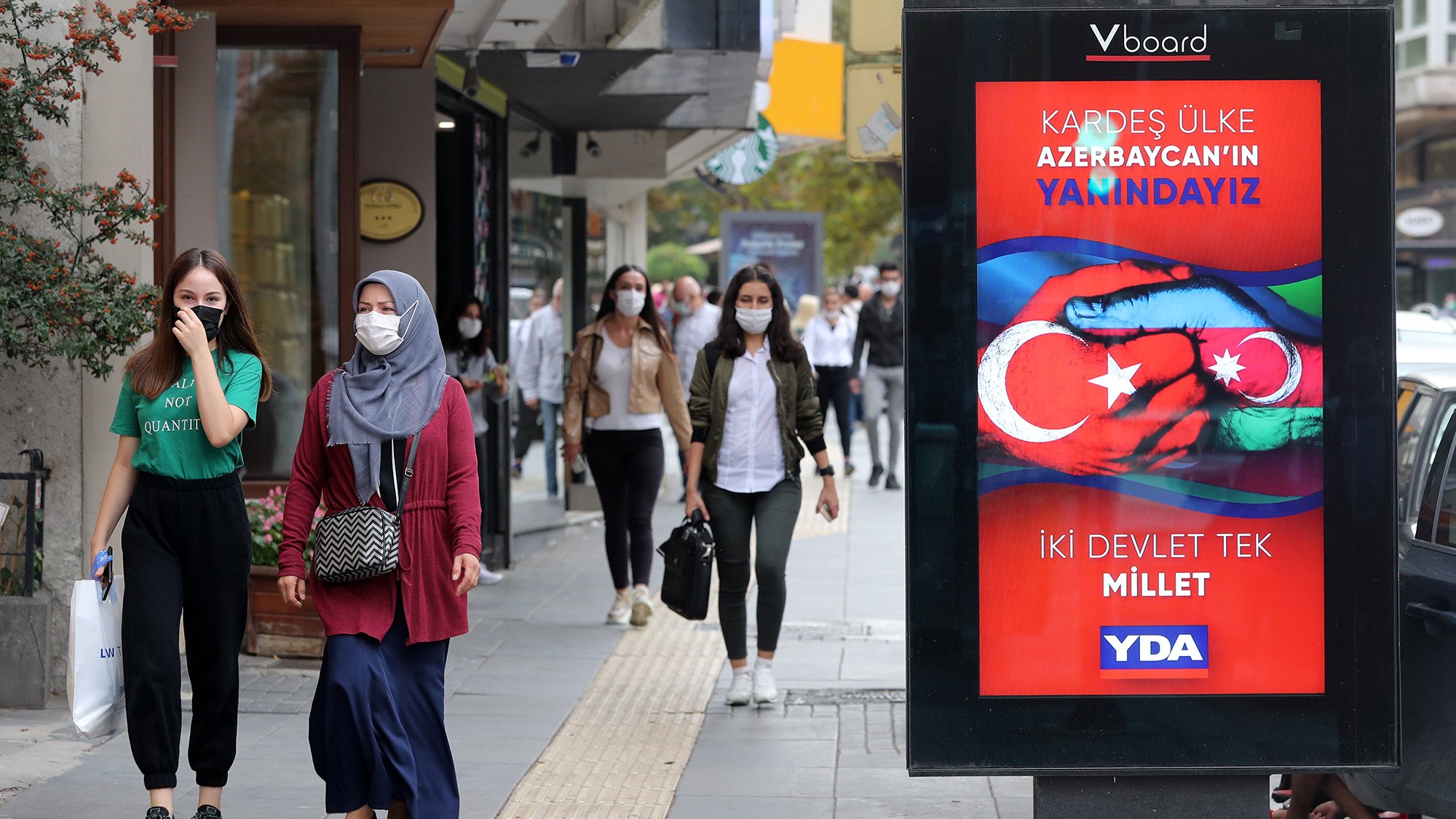 L'image montre une scène de rue animée où des personnes marchent sur un trottoir. À gauche, deux femmes avancent, l'une portant un hijab gris et l'autre en tenue décontractée. À droite, un panneau d'affichage numérique affiche un message en turc, avec des couleurs vives et des symboles représentant les drapeaux de la Turquie et de l'Azerbaïdjan. Le texte semble faire référence à un soutien entre les deux pays. Plusieurs passants peuvent être aperçus en arrière-plan, portant tous des masques de protection.