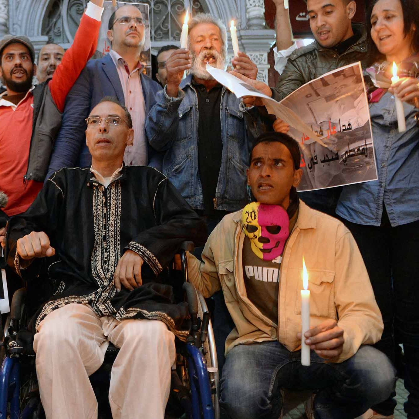 L'immagine mostra un gruppo di persone radunate in un ambiente urbano, apparentemente in un momento di protesta o di vigilanza. Alcuni partecipanti tengono in mano candele accese, mentre altri mostrano cartelli e manifesti con slogan che richiamano il tema della libertà di espressione. Tra le persone c'è un uomo in sedia a rotelle, che sembra essere al centro dell'attenzione. L'atmosfera è di solidarietà e impegno per i diritti civili.