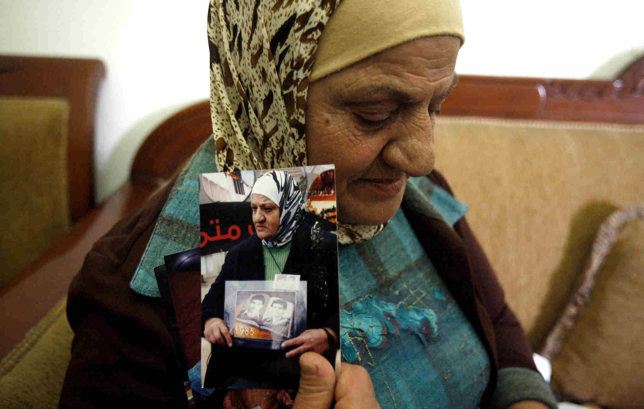 الصورة تظهر سيدة كبيرة في السن ترتدي حجابًا وتجلس على أريكة. تعبر ملامح وجهها عن التأمل والحنين، حيث تُمسك بعدد من الصور الفوتوغرافية في يديها. يبدو أن هذه الصور تحمل ذكريات مهمة بالنسبة لها، وقد تكون تتعلق بأفراد من عائلتها. الخلفية بسيطة وتُشير إلى بيئة منزلية مريحة.