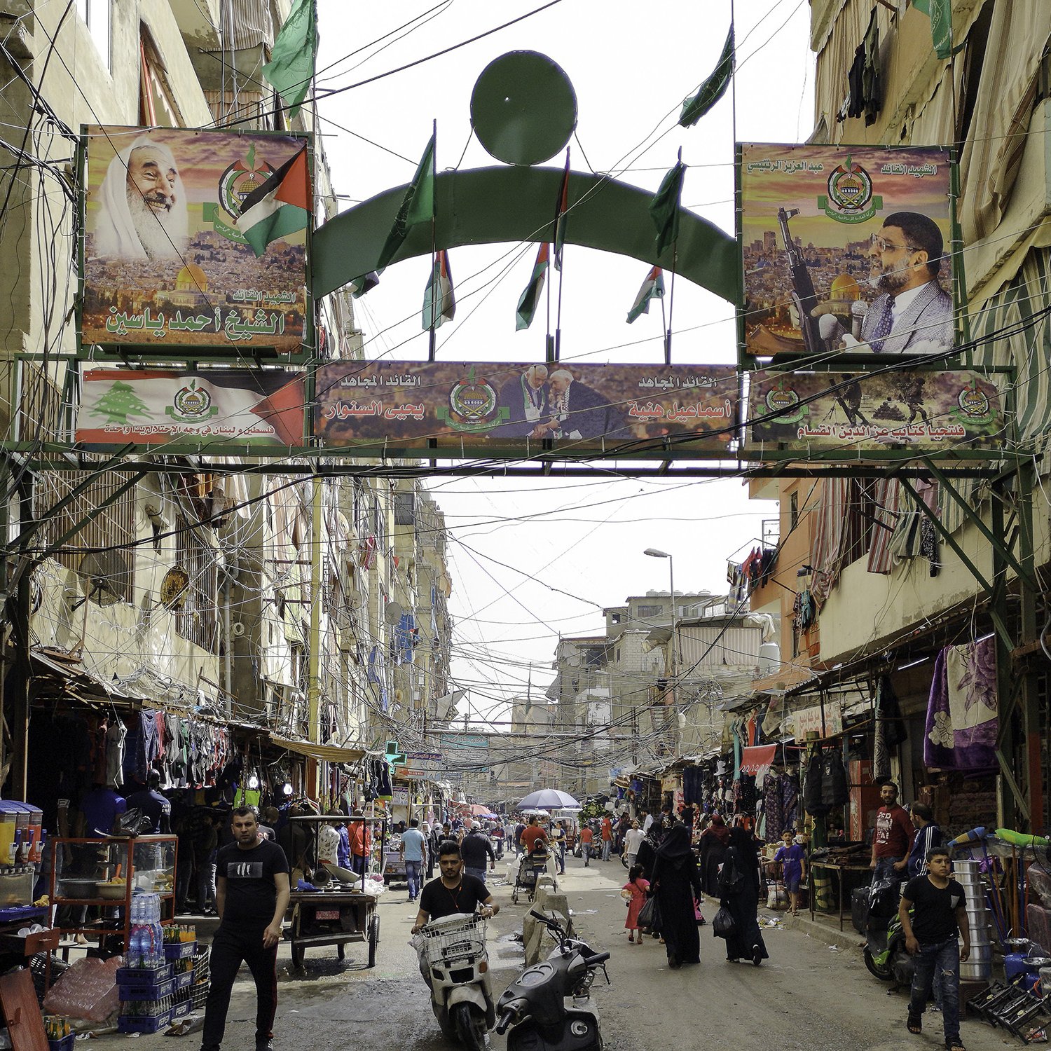 L'image montre une rue animée d'un quartier urbain. On peut y voir des bâtiments étroits et des fils électriques visibles. Des drapeaux et des affiches, probablement liés à un événement ou à une célébration, décorent l'entrée de la rue. Des personnes circulent, certaines à pied et d'autres à moto, ajoutant à l'activité de la scène. Le lieu semble être un marché ou un centre commercial, avec des étals vendant divers produits. L'atmosphère est vibrante et vivante.