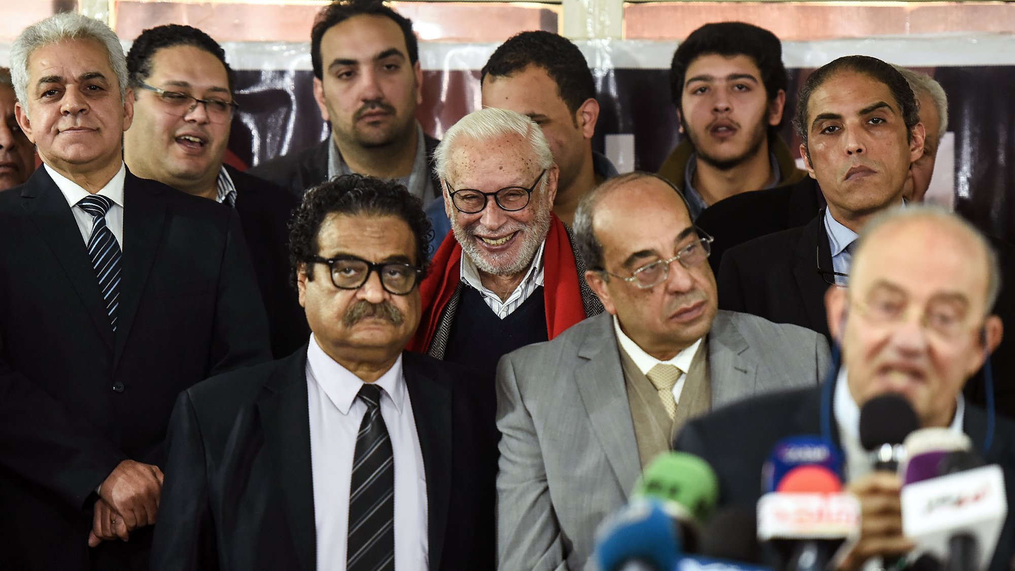 این تصویر گروهی از مردان را نشان می‌دهد که در یک رویداد یا مراسم جمع شده‌اند. در مرکز تصویر، یک مرد با موهای سفید و لبخند بزرگ دیده می‌شود. دیگران به او نزدیک هستند و به صحبت‌های شخصی که در حال ارائه سخنرانی است، گوش می‌دهند. افراد لباس‌های رسمی پوشیده‌اند و برخی از آن‌ها مشغول به صحبت یا تبادل نظر هستند. محیط تصویر به نظر می‌رسد به یک مکان رسمی یا اداری تعلق دارد.