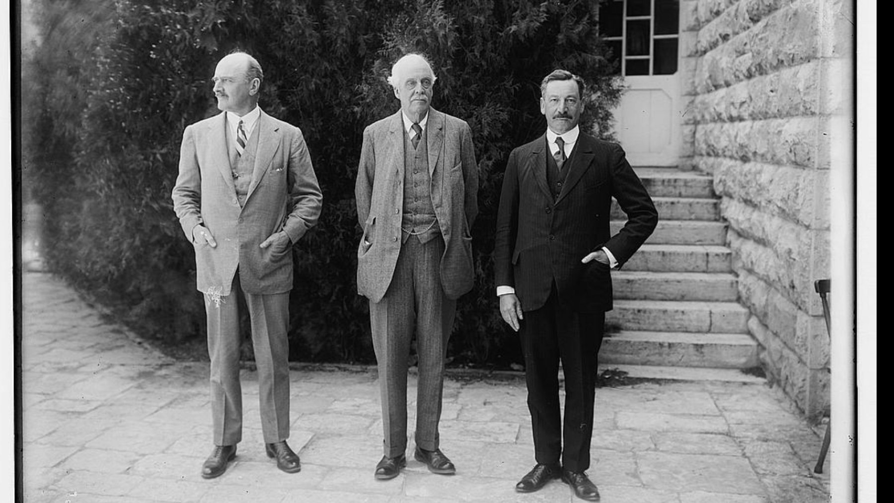 سه مرد در لباس رسمی ایستاده‌اند. پس‌زمینه شامل درختان و یک سازه سنگی ساده است.